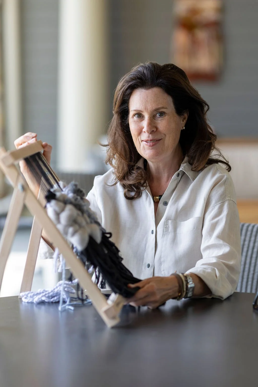 Woman sitting at a table holding a yarn sample board, smiling, with a loom in front of her.