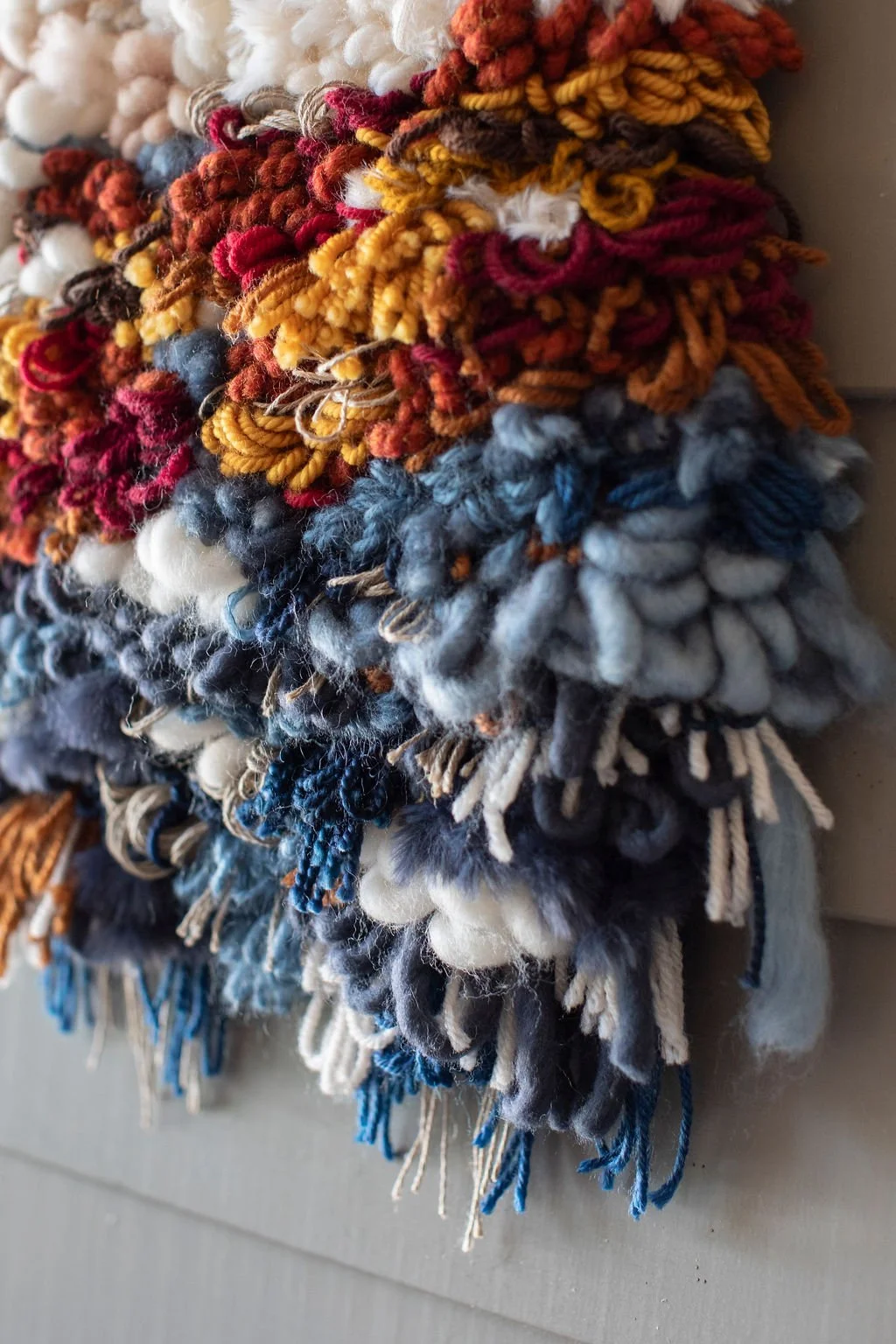 Close-up of colorful, textured yarns and wool rug with shaggy, knotted loops in multiple shades including blue, gray, black, white, red, orange, yellow, and beige.