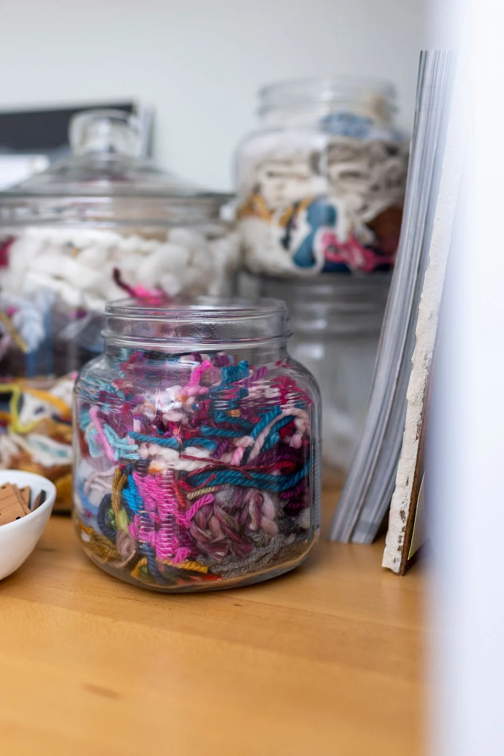 A clear glass jar filled with colorful yarn balls sitting on a wooden surface, with other jars and supplies in the background.