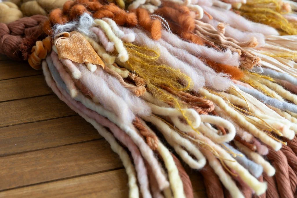 Close-up of a colorful, textured woven tapestry made from various yarns and fibers, resting on a wooden surface.