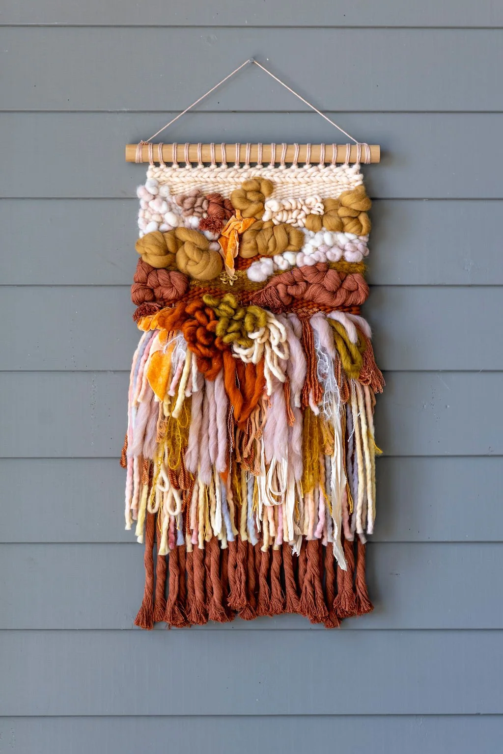Colorful woven wall hanging with various yarn textures and shades, hanging on a gray wall.
