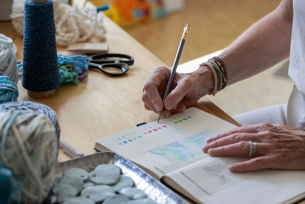 A person with bracelets on their wrist is writing or drawing in a sketchbook with a pencil, sitting at a table with yarn, scissors, and other supplies.