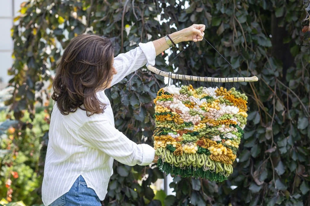 Woman holding a colorful, textured woven art piece on a wooden hanger outdoors next to a large leafy bush.