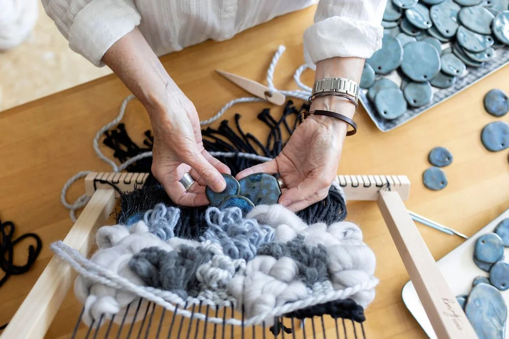 A person weaving a textile with blue, white, and black yarn using a small loom, with blue oval-shaped stones on a tray and table nearby.