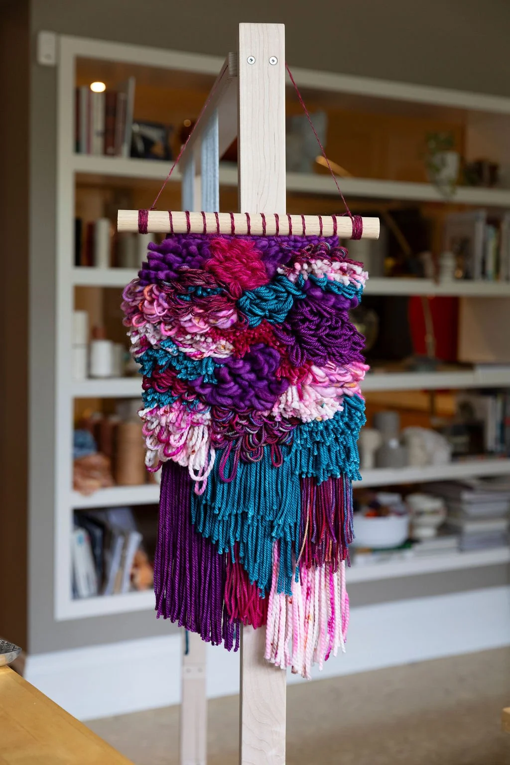 Colorful yarn wall hanging with shades of purple, pink, blue, and white, hanging on a wooden frame indoors with bookshelves in the background.