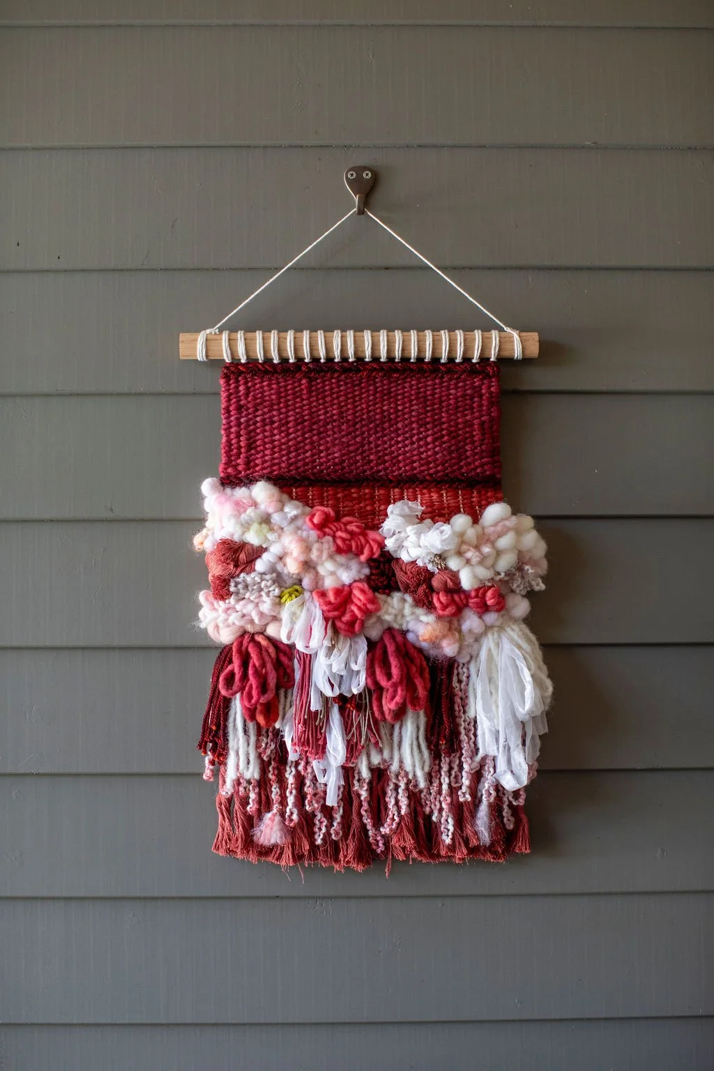 Colorful, textured wall hanging with red, pink, and white yarns, tassels, and pom-poms, hanging on a gray wooden wall.