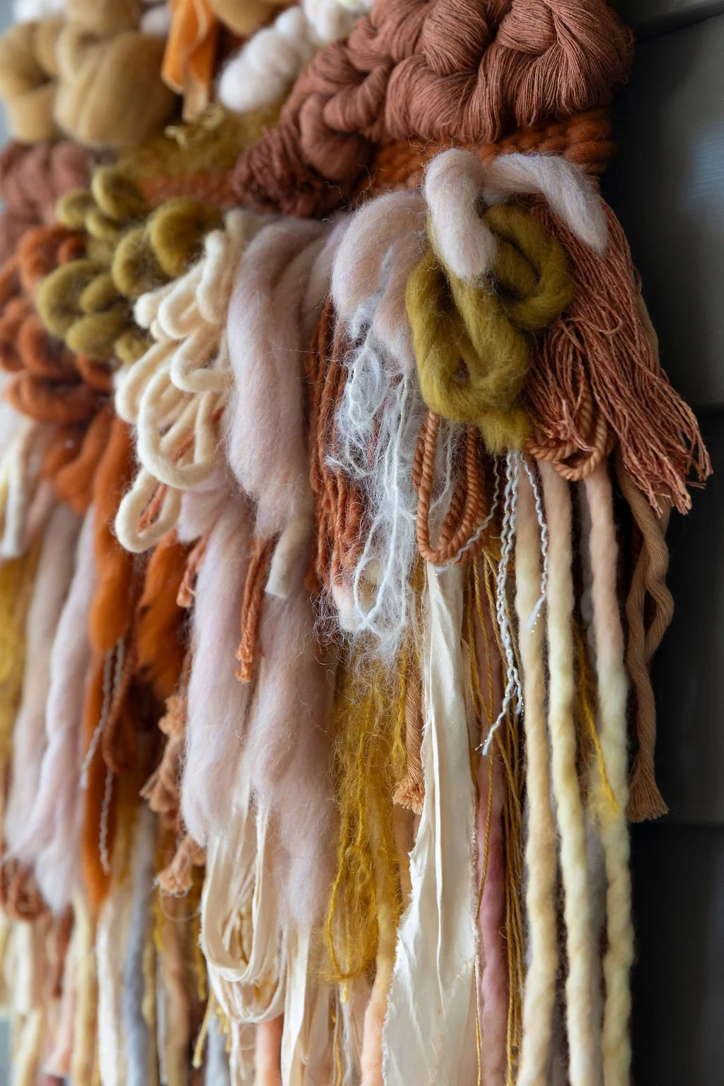 Close-up of woven textile art made from multicolored wool yarns, featuring fringes and knots in warm earthy tones of beige, cream, orange, brown, and mustard yellow.