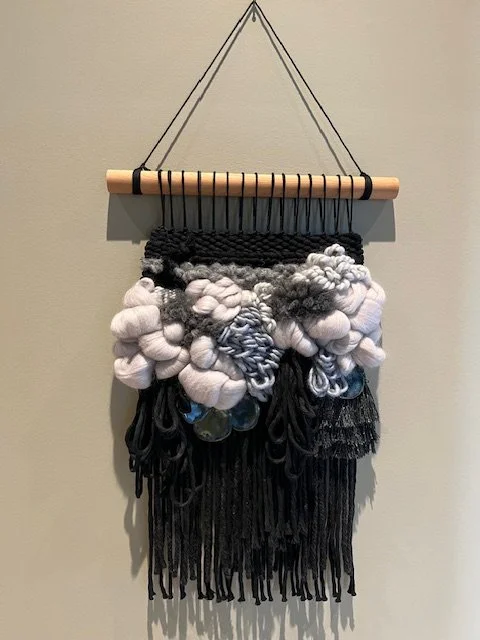 A wall hanging made of yarn and fabric knots, suspended from a wooden dowel with black string.