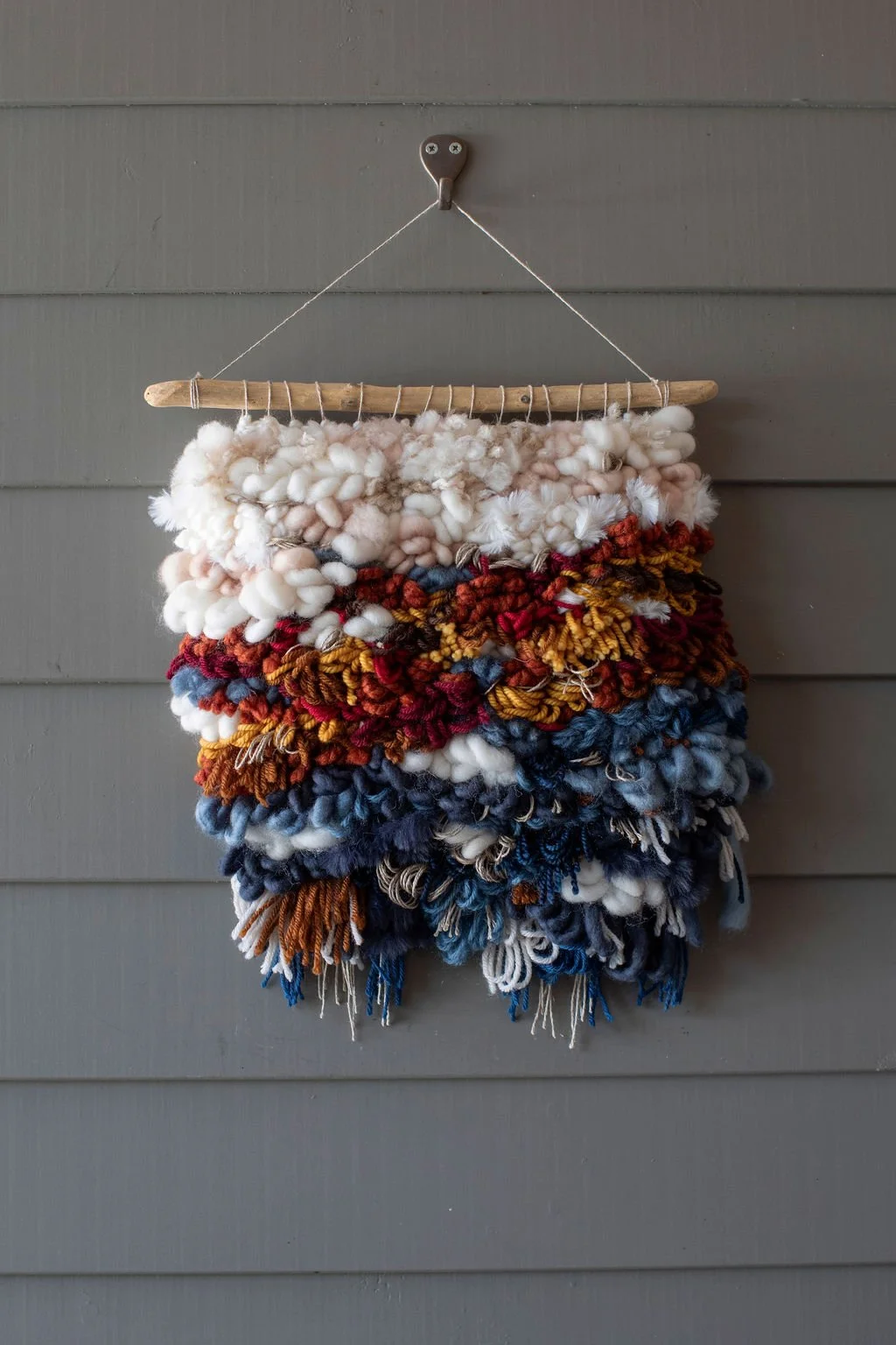 Colorful woven wall hanging made of chunky yarn, hanging from a wooden rod on a gray wall.