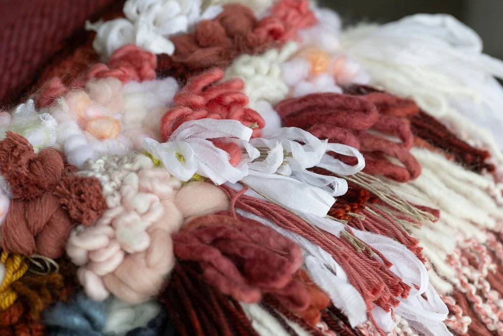 Close-up of a colorful woven textile with various textured yarns, ribbons, and fabric strips in shades of red, white, pink, and beige.