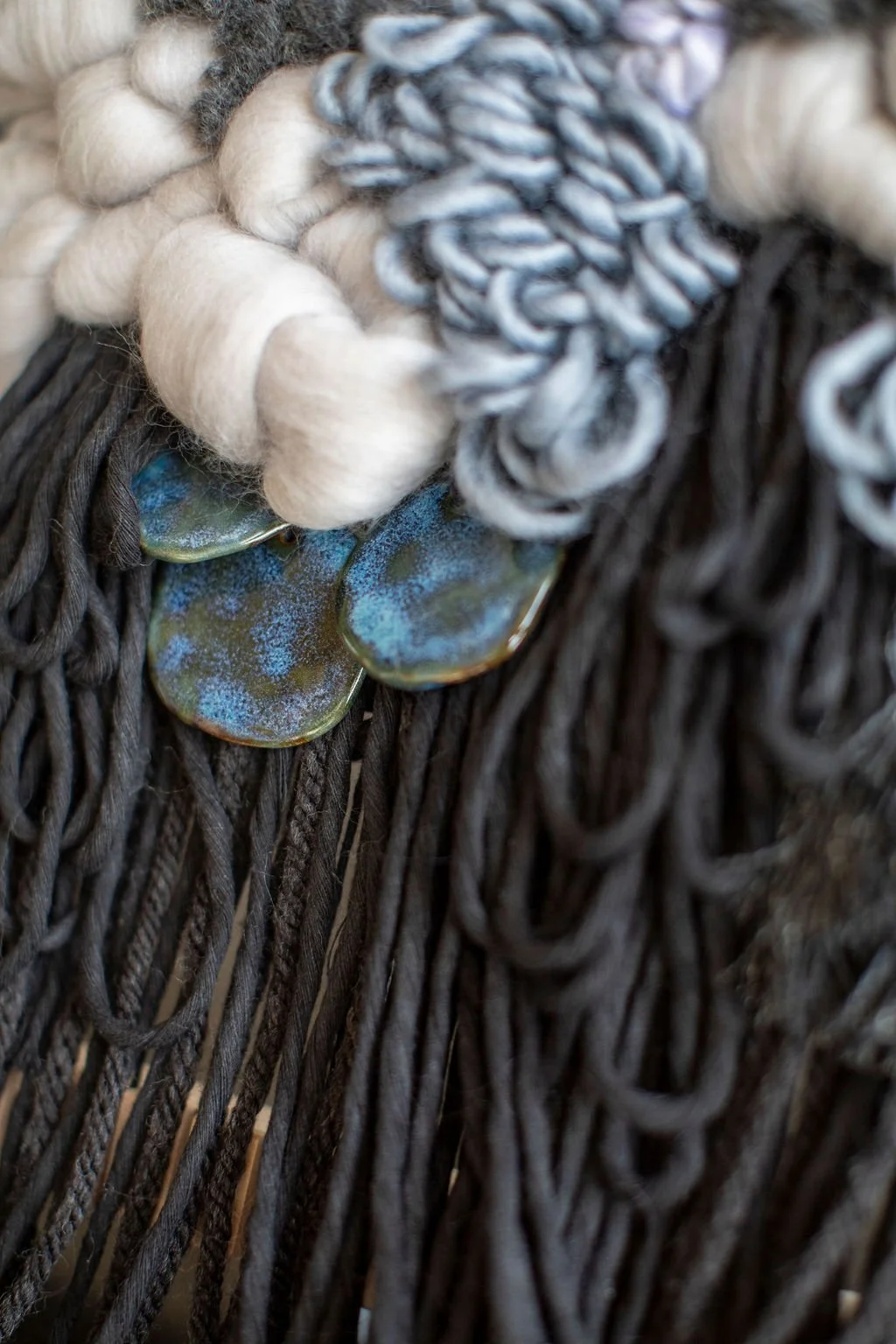 Close-up of a woven decorative piece consisting of dark brown ropes, with white and black thick yarns and a string of decorative metal discs with iridescent finish.