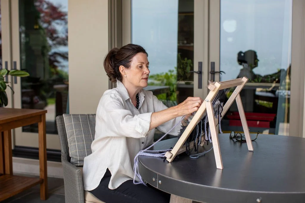 A woman sitting at a table, knitting on a small wooden loom.