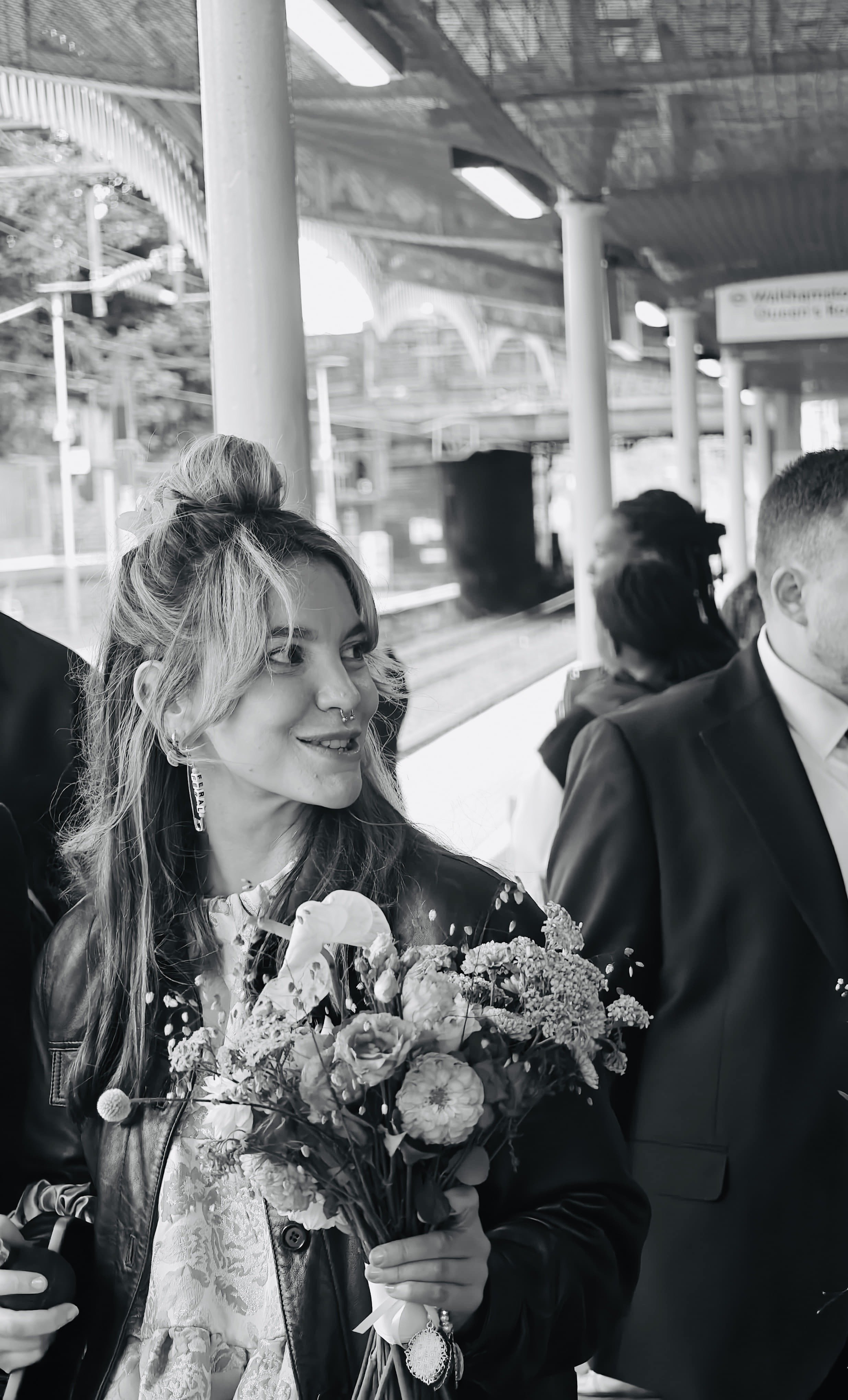 Black and white image of a blonde woman with a septum piercing, long earrings, hair in half up, half down style, leather jacket, holding a bouquet of wedding flowers. She is at a train station with groomsmen just out of shot.