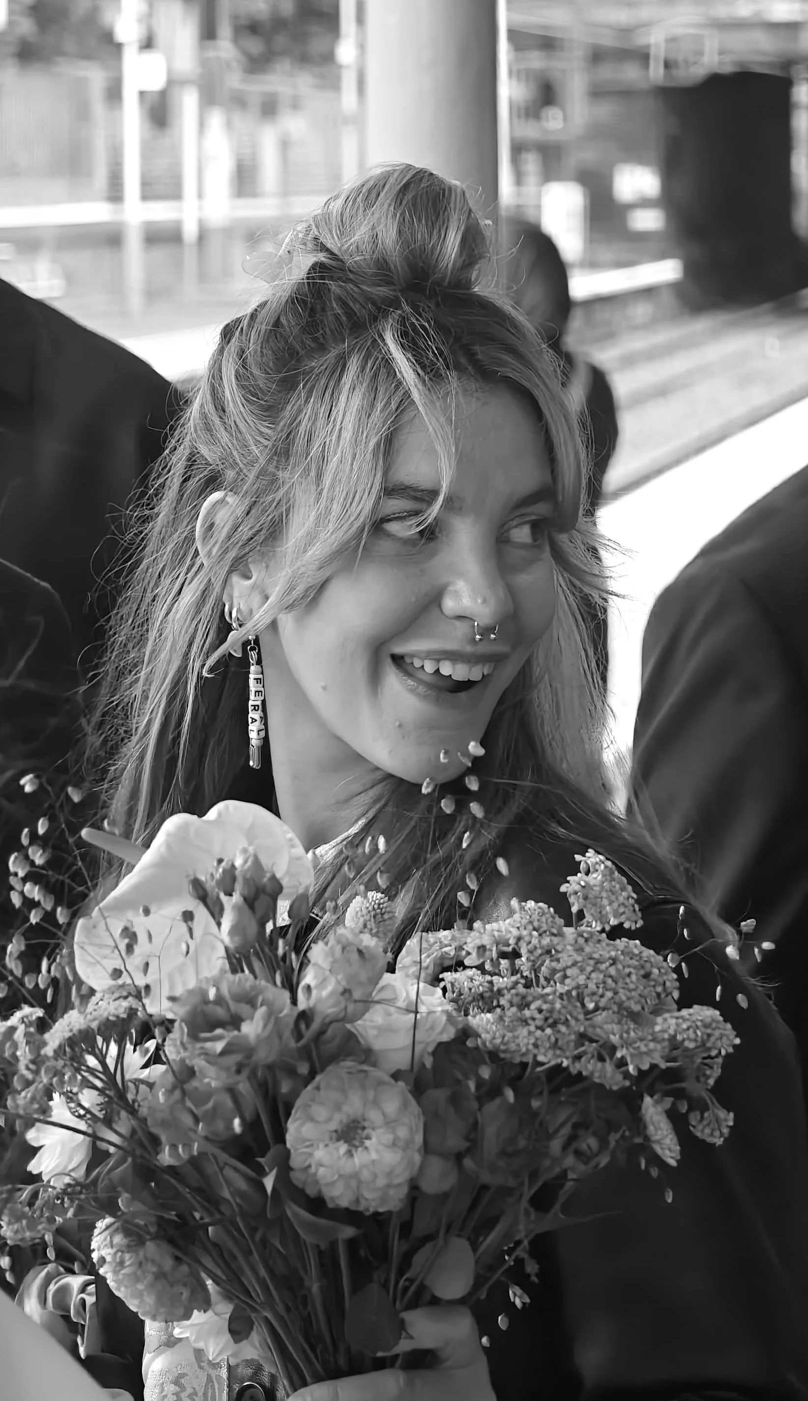 Black and white image of a blonde woman with a septum ring and earrings reading "Feral", holding a bouquet, looking over her shoulder mischievously