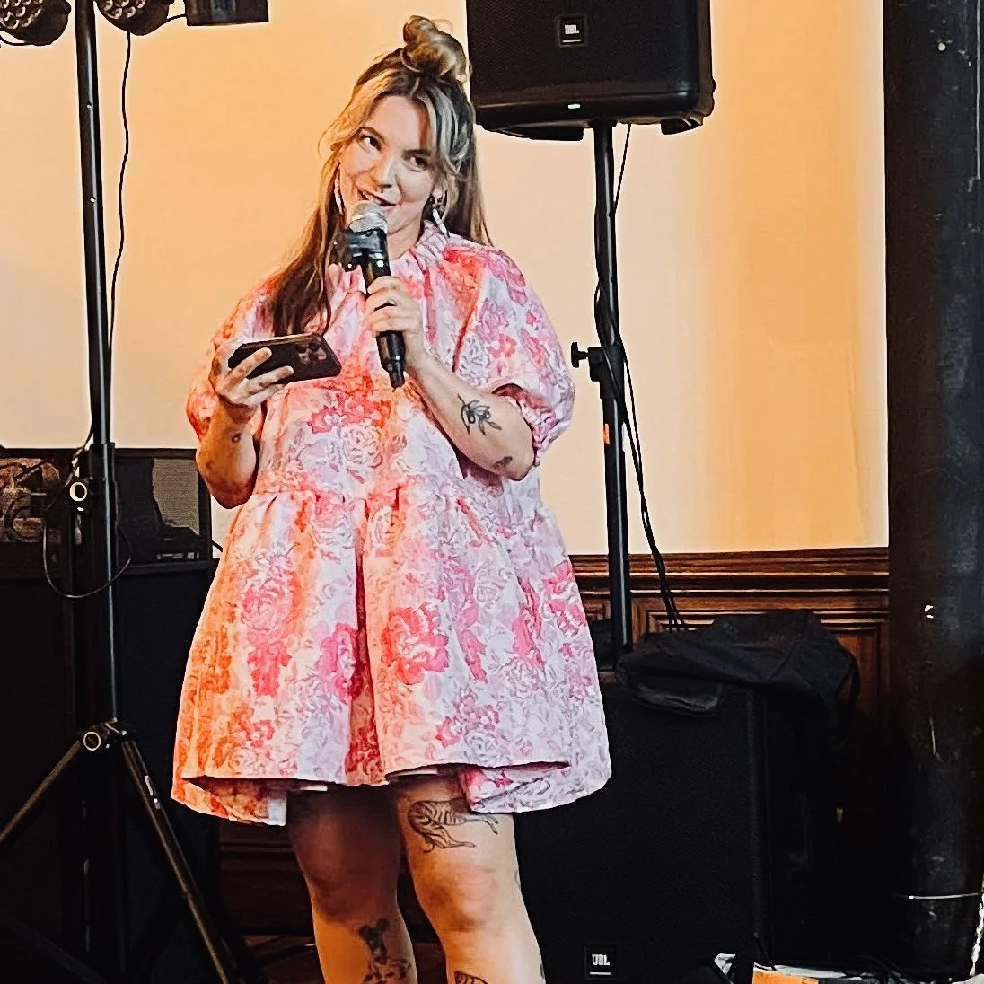 Blonde woman in a floral pink dress, with tattoos on her arms, holding a microphone and presenting to an audience. Yellow background and speakers can be seen.