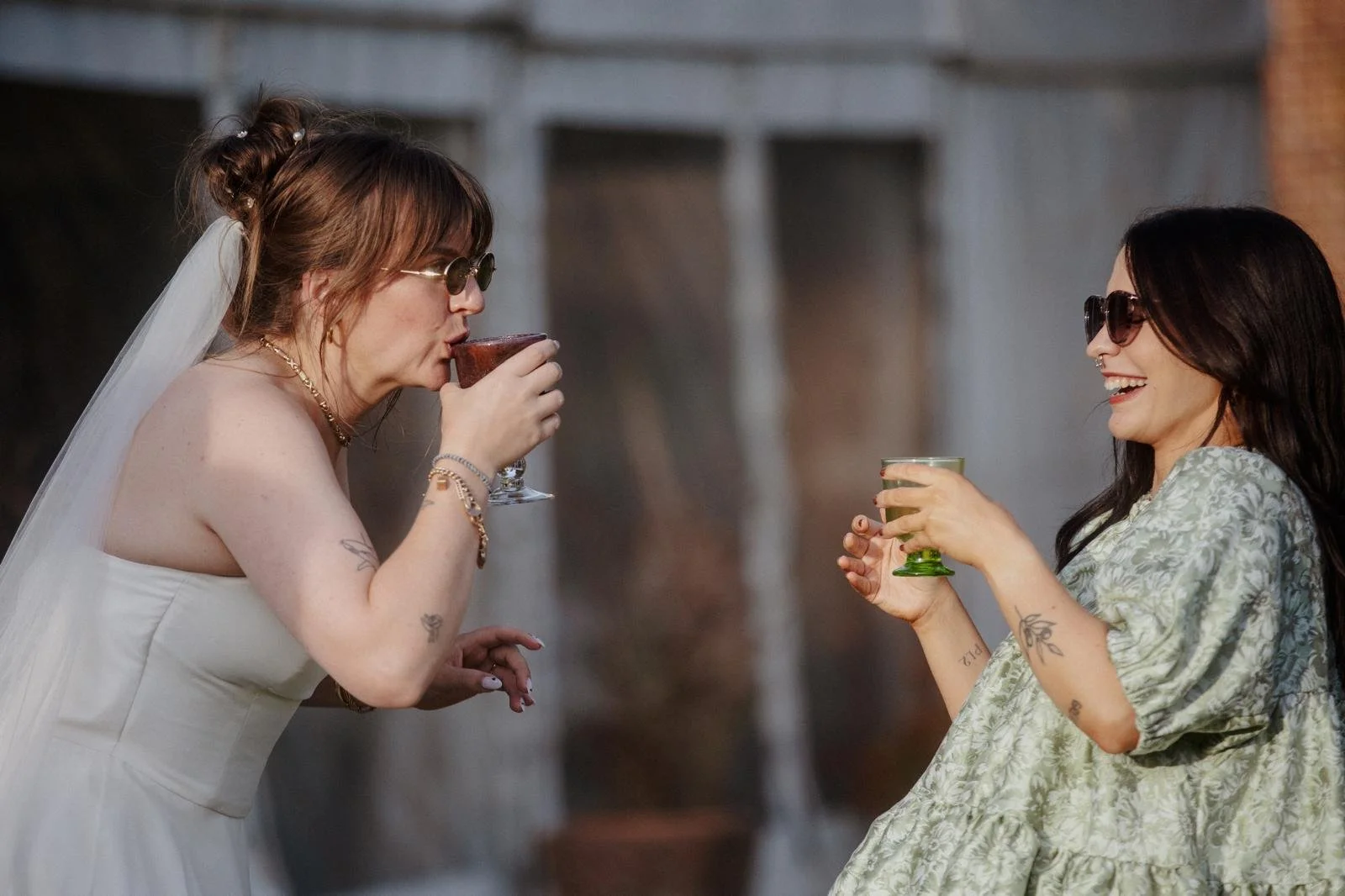 Colour image of a bride in a strapless ivory dress, gold jewellery, veil and sunglasses with light brown hair in a casual up-do, sipping a drink. Also pictured is a woman with long dark hair, sunglasses, a septum ring and a green dress, laughing.