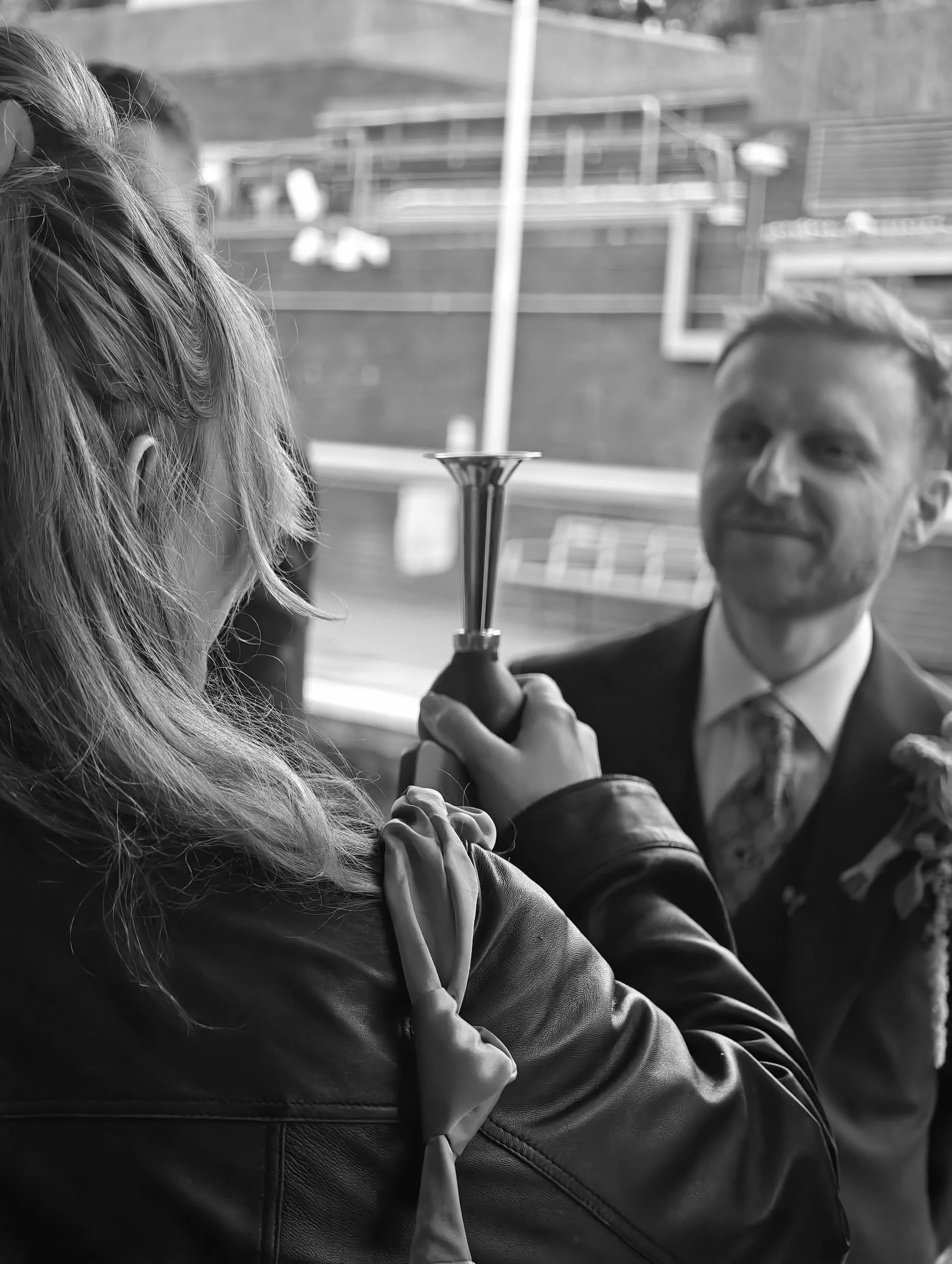 Black and white close up image of a woman with blonde hair holding a bike horn. Groom blurred in the background smiling