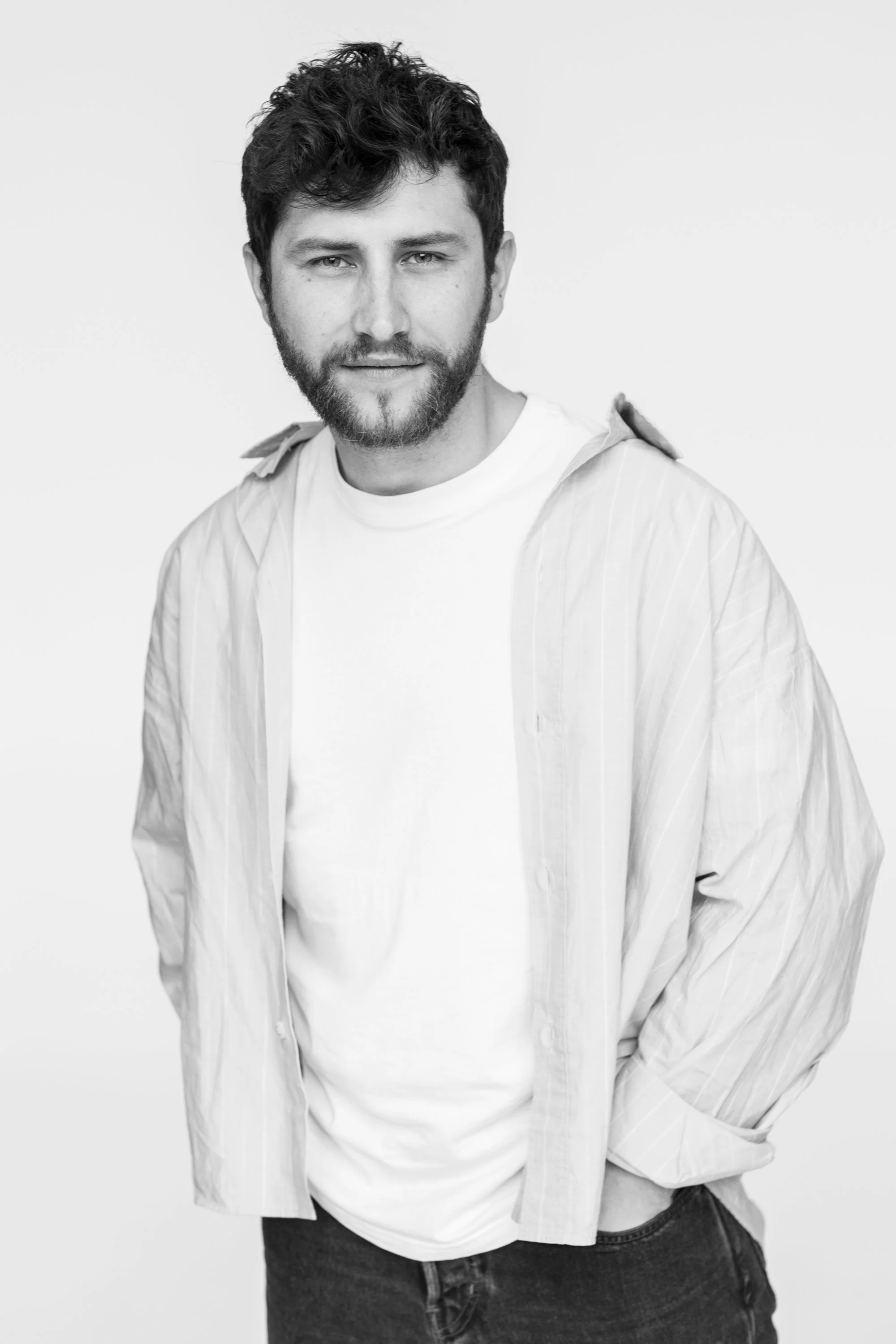 Black and white portrait of a young man with curly hair, beard, and mustache, wearing a white shirt with rolled-up sleeves and dark jeans, standing against a plain light background.