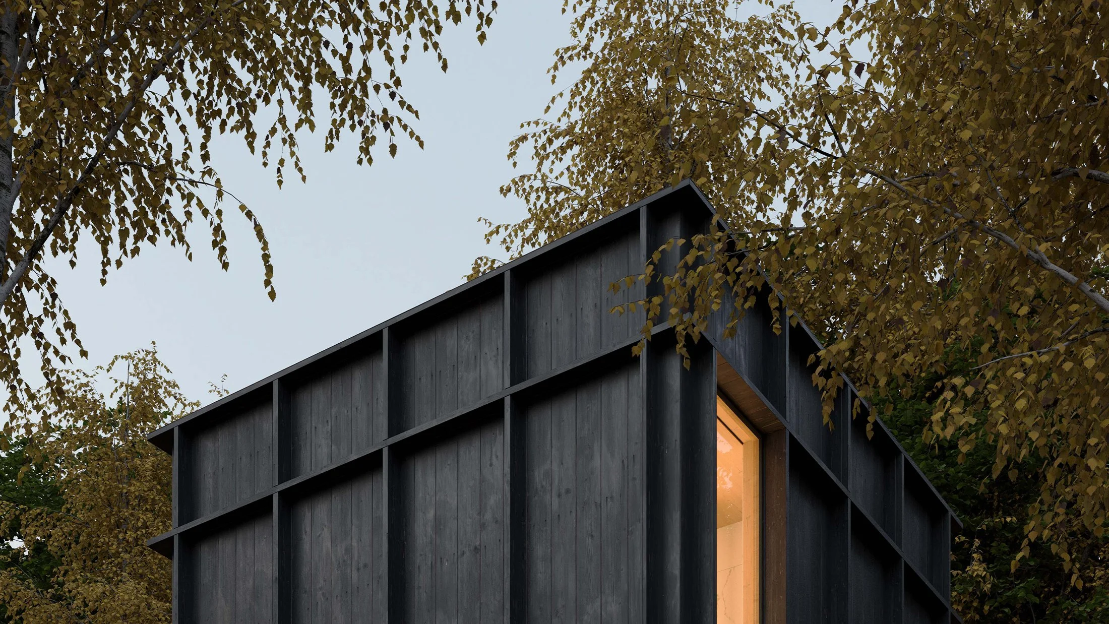 A modern black wooden house with a large window revealing the warm interior light, surrounded by trees with golden autumn leaves, under a partly cloudy sky.