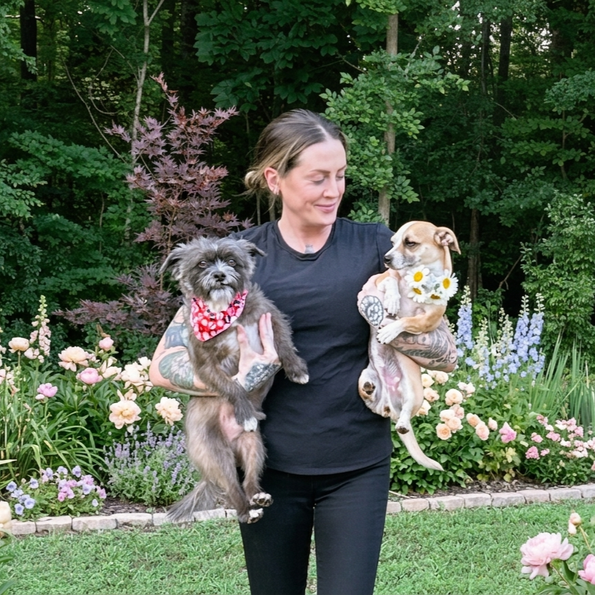 Woman holding two dogs in a garden with colorful flowers and green trees.