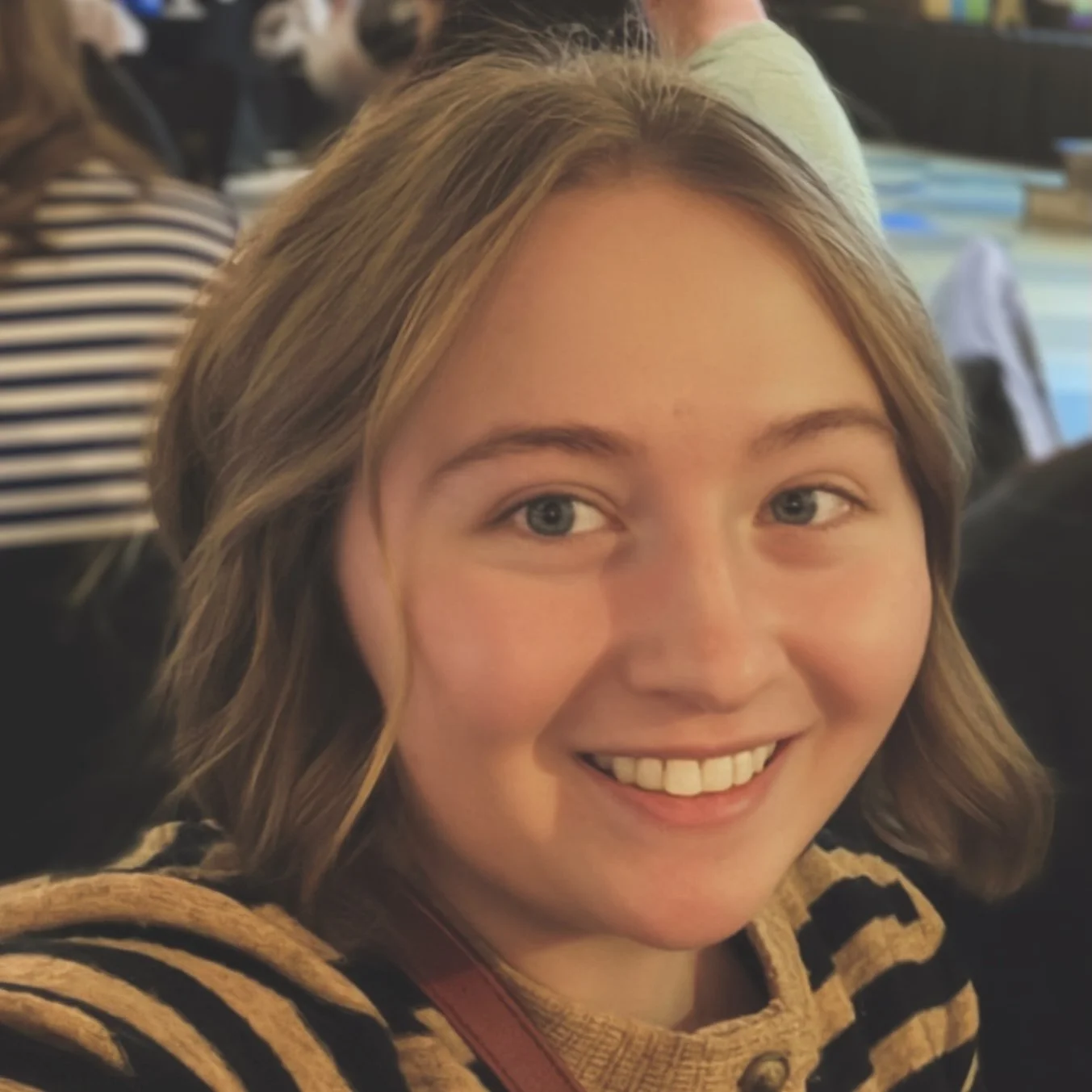 A young woman with wavy blonde hair and blue eyes smiling, wearing a black and yellow striped sweater, in an indoor setting with other people and books in the background.