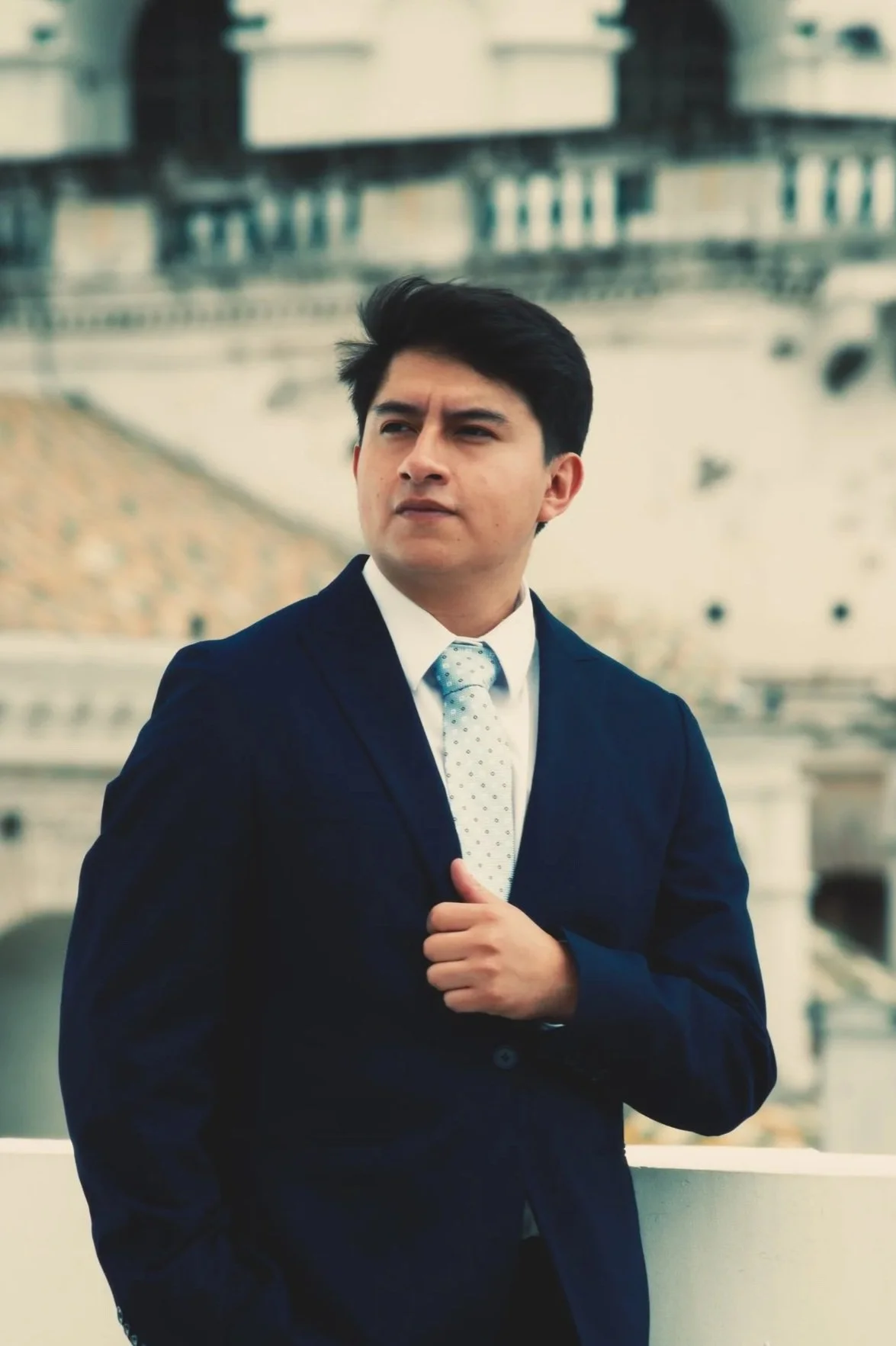 Un abogado con traje oscuro, camisa blanca y corbata de puntos, de cabello oscuro, de fondo una ciudad con edificios y puentes.