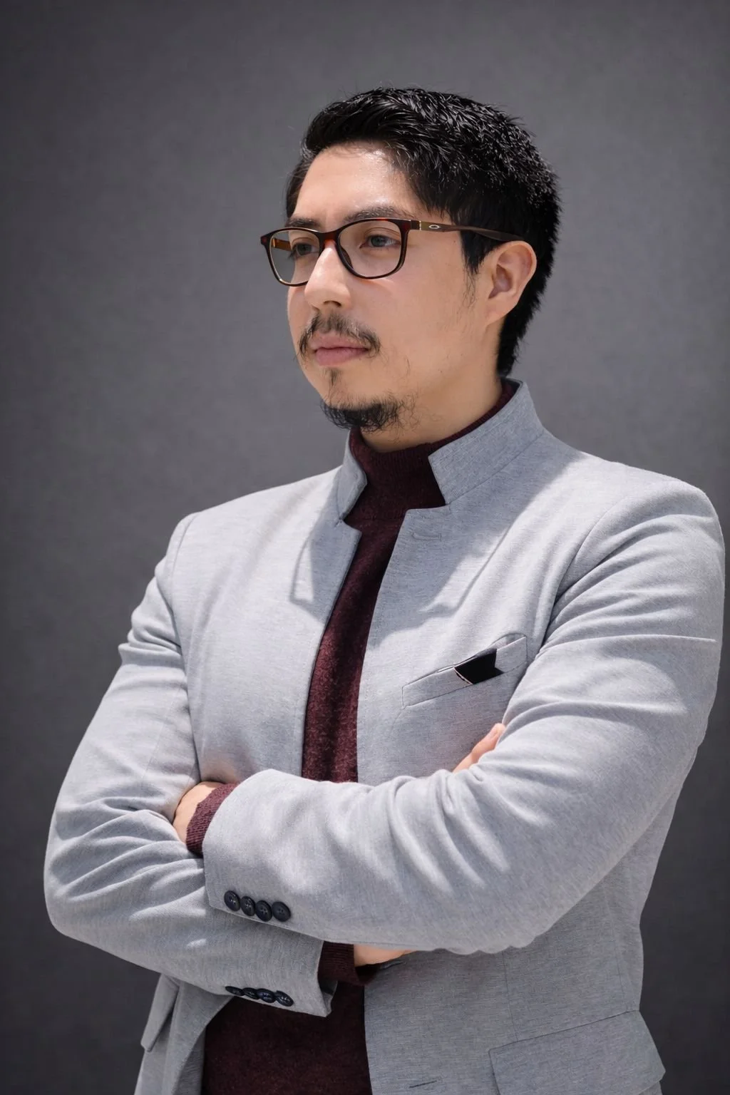 Un Abogado con gafas, barba y bigote, vestimenta formal gris con una camisa de cuello alto burdeos, de cabello oscuro, con los brazos cruzados, en un fondo gris oscuro.