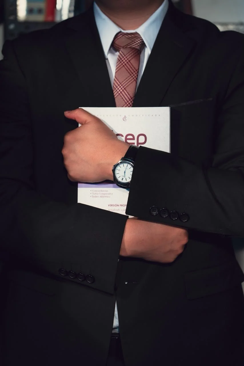 Hombre con traje negro, camisa blanca, corbata a cuadros, sosteniendo una carpeta con un libro o documento, lleva un reloj en la muñeca izquierda, en un entorno urbano.