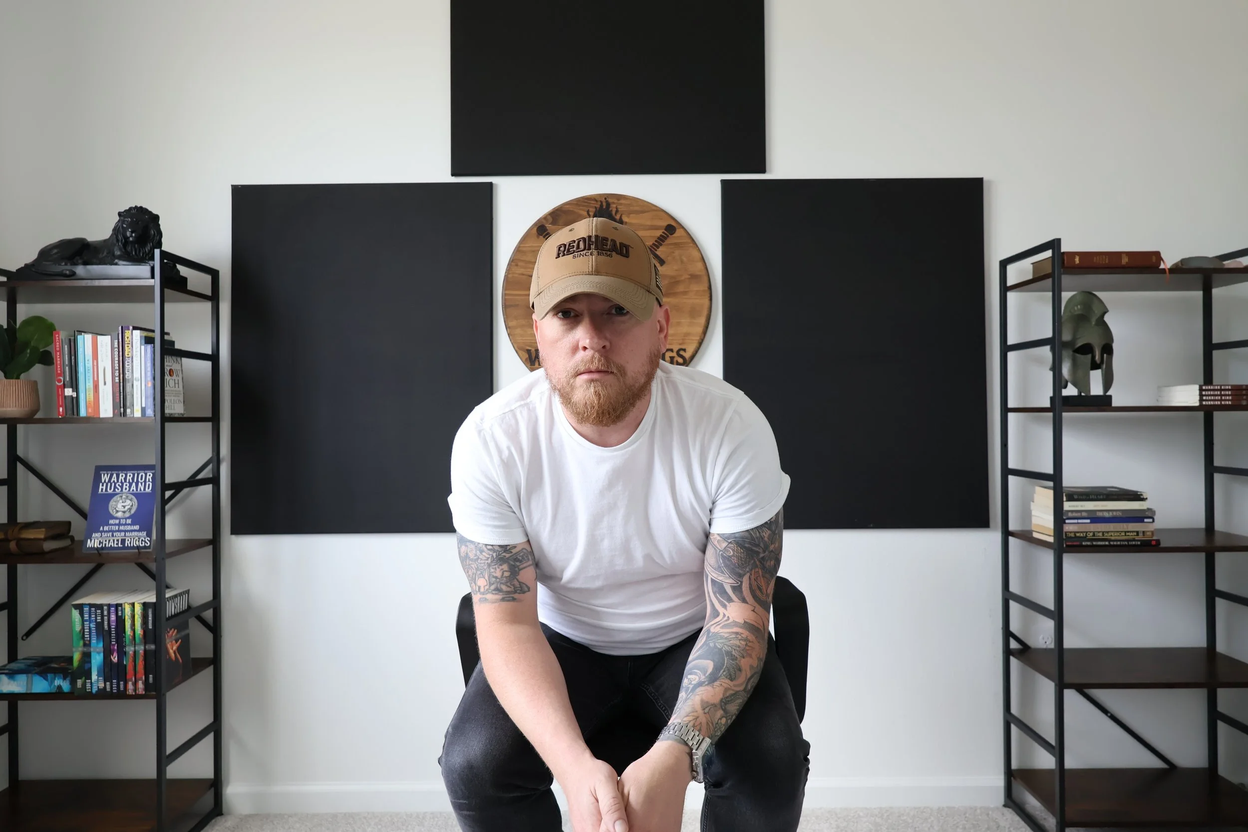 A man with tattoos on his arms, wearing a tan baseball cap with redhead logo, white t-shirt, and black pants, is sitting in a modern room with bookshelves, black and wood decorative elements, and black wall panels.