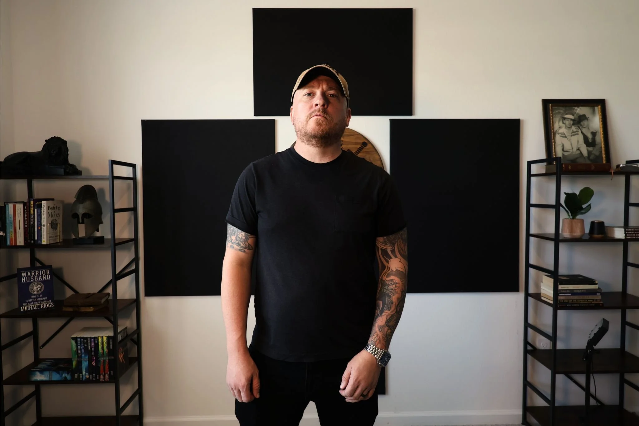 Man with tattoos wearing a black T-shirt and cap standing in front of a decorative wall with black panels and bookshelves with various items, including a framed black-and-white photo and a face sculpture.