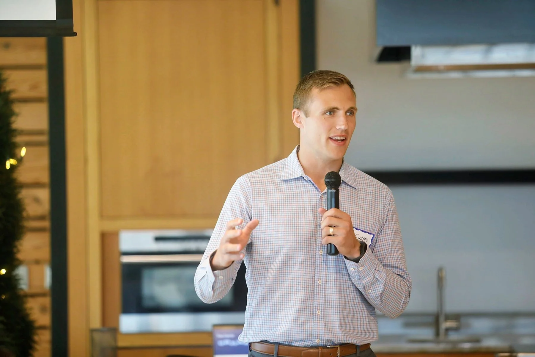 A young man with short blond hair, wearing a light blue checkered shirt and brown belt, is speaking into a microphone during a presentation or speech in a room with wooden and metal elements.