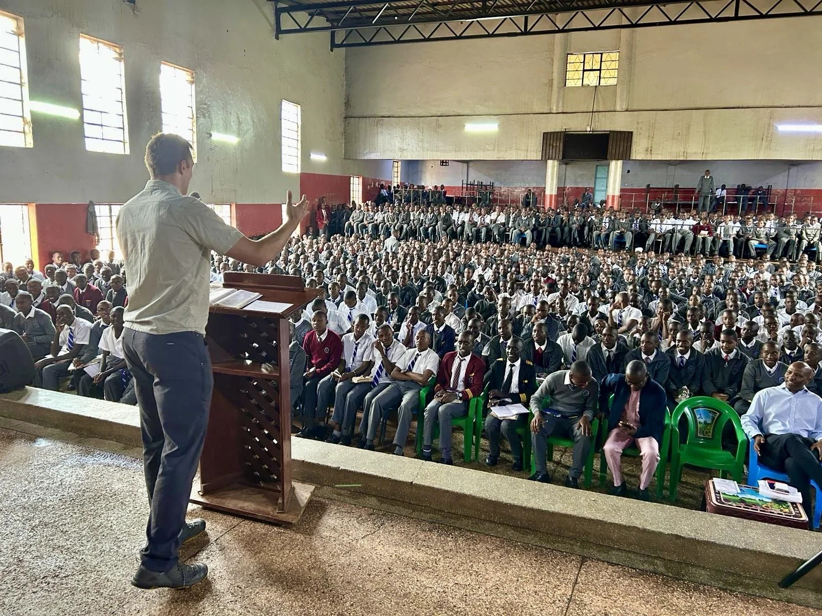 A man giving a speech at a podium in a large classroom or hall filled with students, some sitting in the front and others on elevated seating in the back, with sunlight coming through windows.