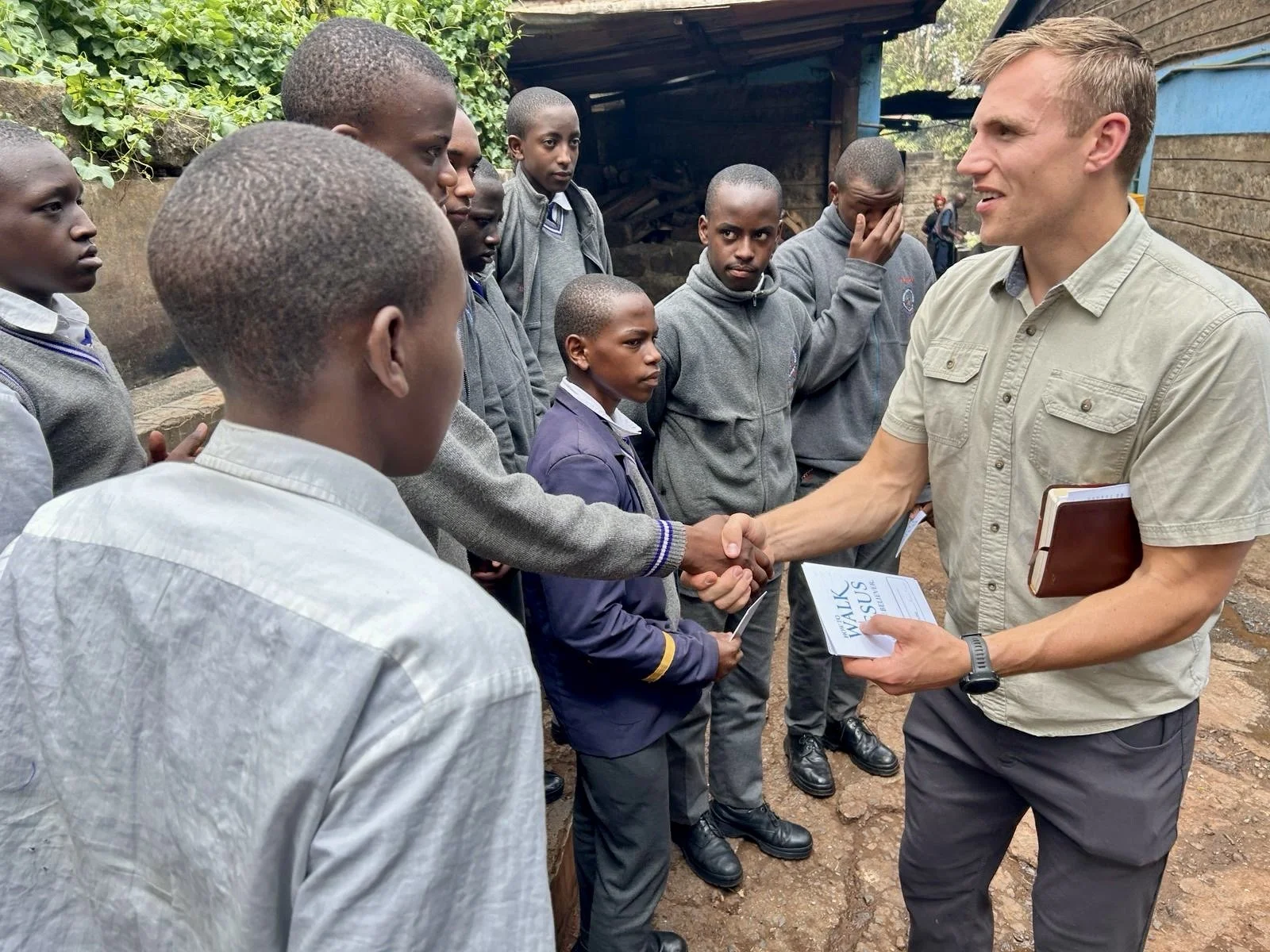 A man with light-colored hair and a beige shirt is shaking hands with a group of students outside. He is holding a notebook and a pamphlet that says 'WALK 2 MOSUL'. The students are wearing school uniforms and standing in a line, some looking serious or distracted. The background shows a wooden structure and greenery.