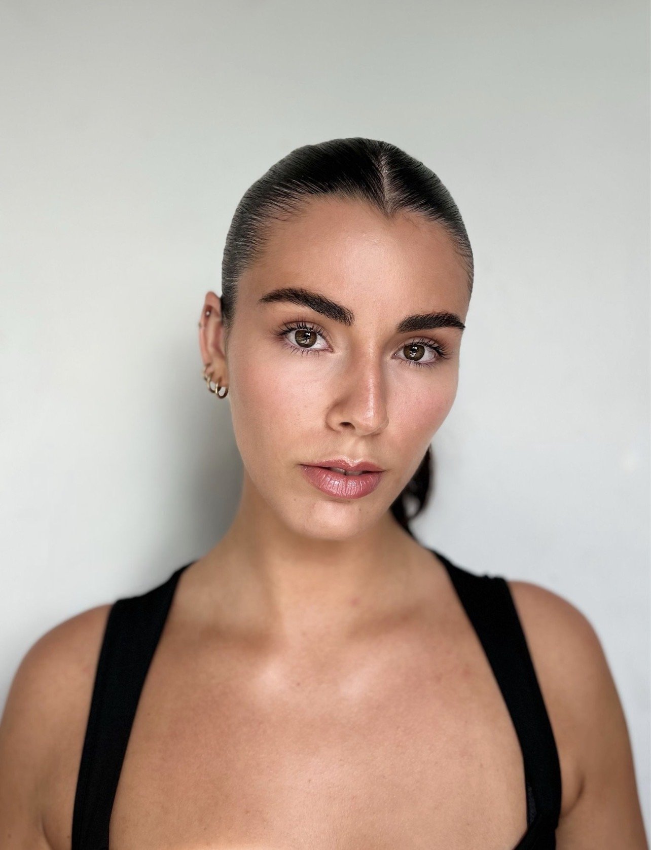 Retrato de uma mulher com pele clara, cabelos escuros presos para trás, sobrancelhas grossas, olhos claros, usando brincos pequenos e um top preto, contra fundo branco.