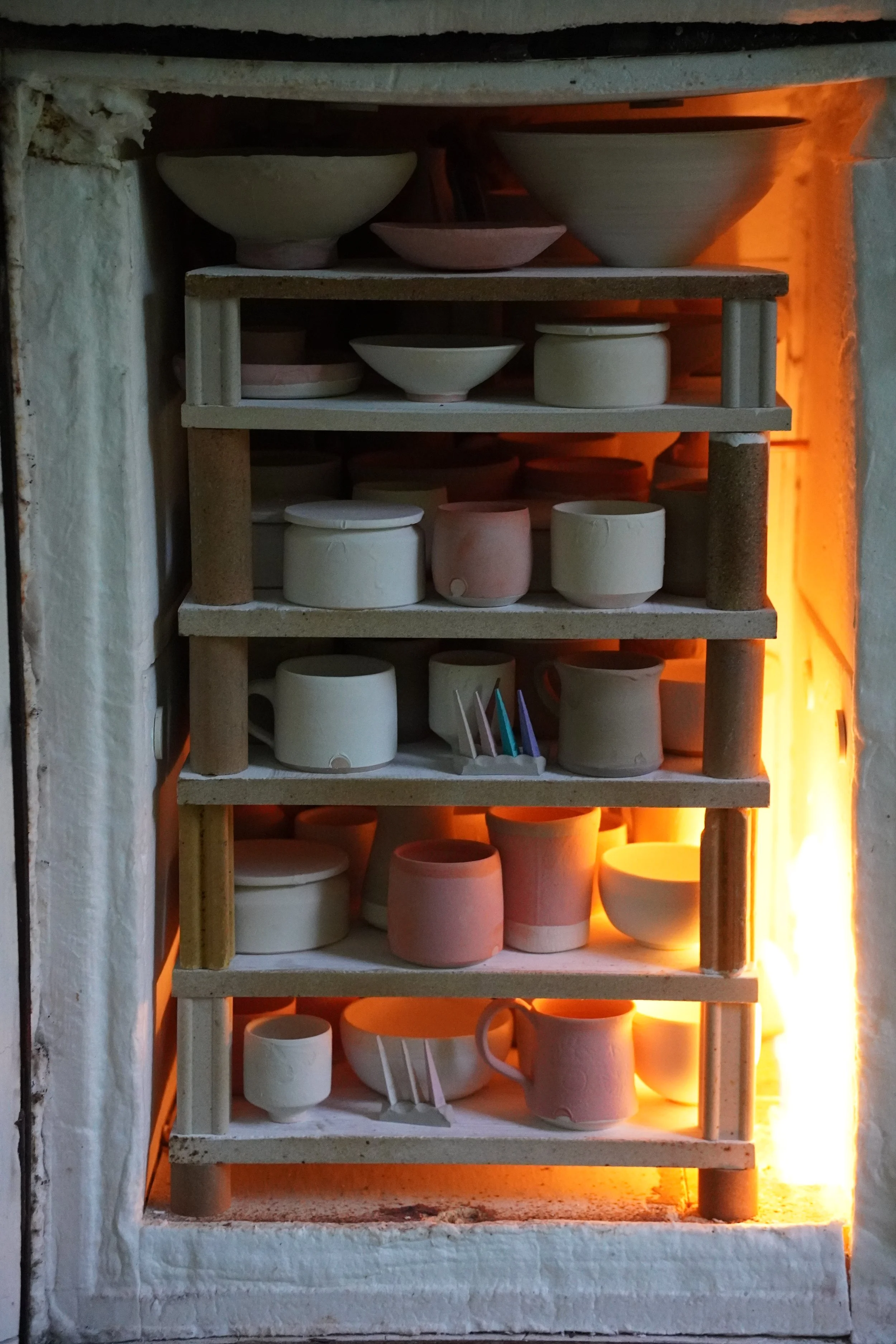 A pottery kiln with multiple shelves containing various ceramic bowls and cups, glowing orange from the heat inside.
