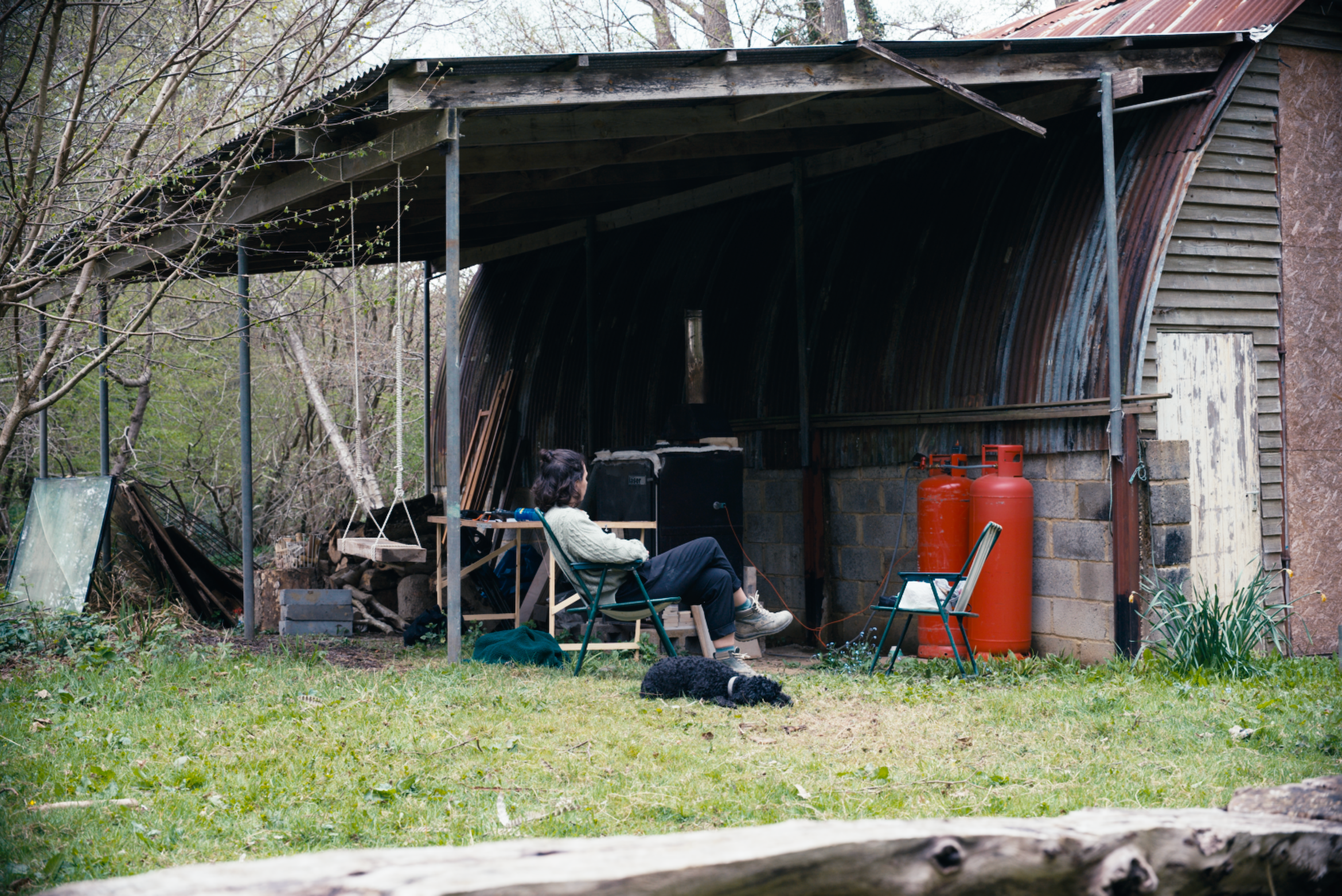 A woman sitting on a lawn chair, with a dog lying on the grass nearby the kiln site, in a wooded backyard setting with trees and a rustic shed.