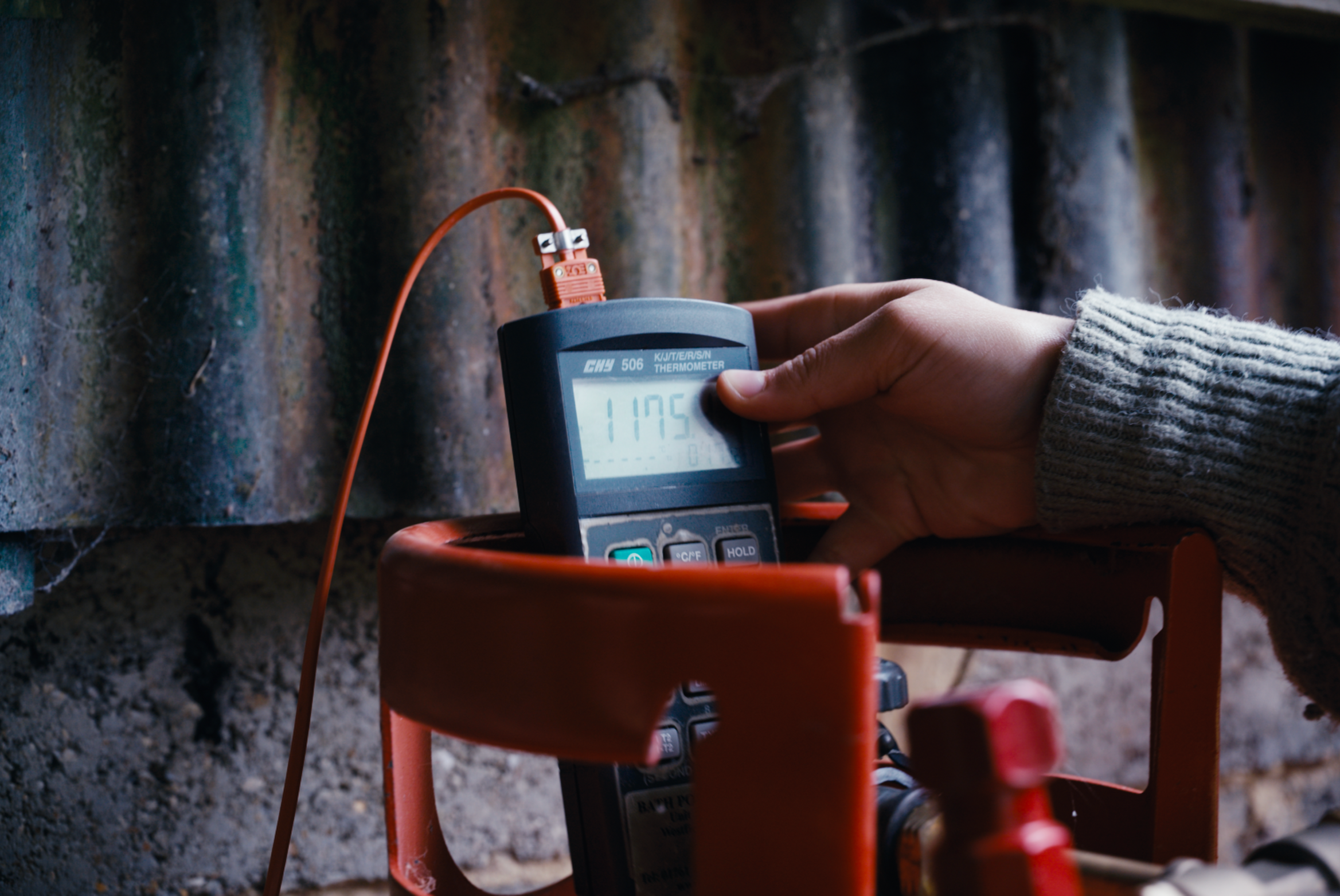 A person holding a digital thermometer measuring 117.5°F near a wall with moss and small waterfalls.