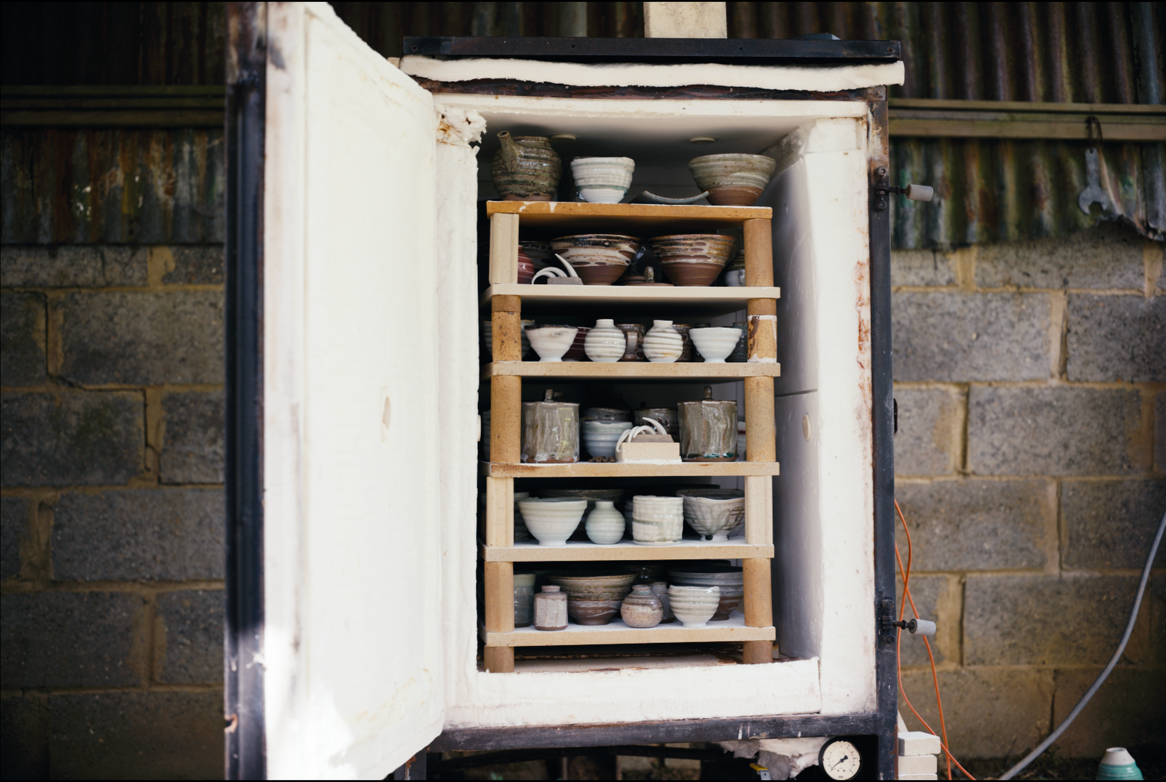 Open kiln with multiple shelves of ceramic bowls and pots, surrounded by stone and metal structure.