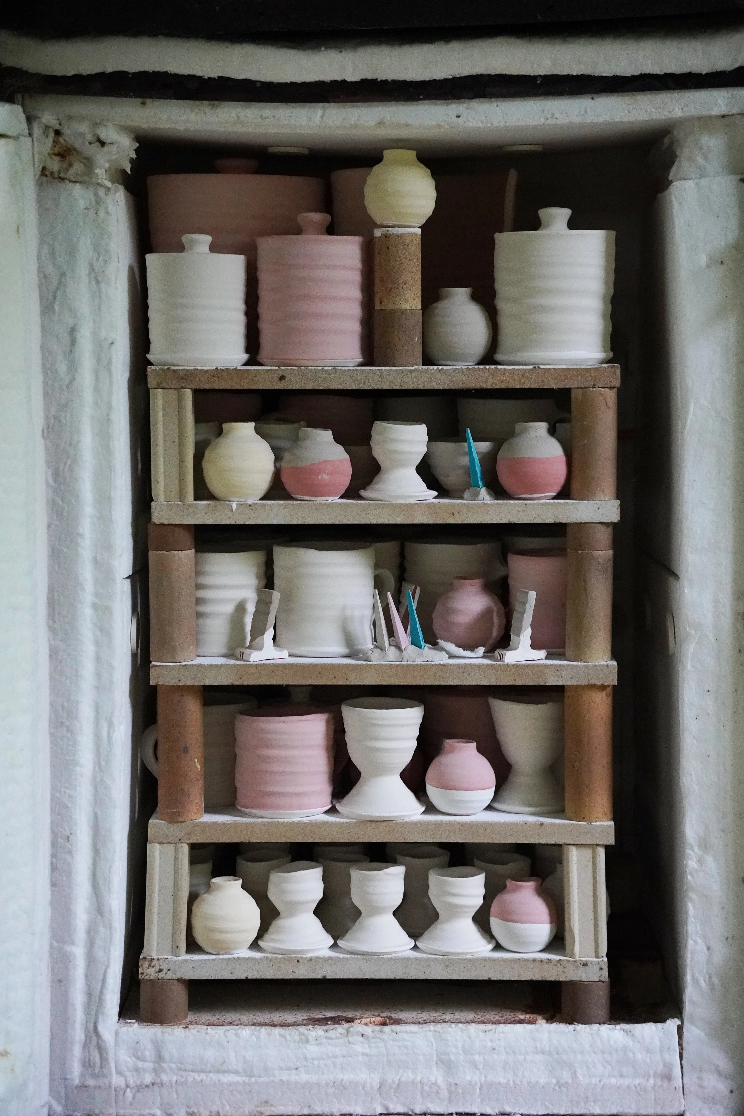 Shelves with unglazed ceramic vases and pots, some with pink or cream coloring, displayed in a pottery studio or shop.