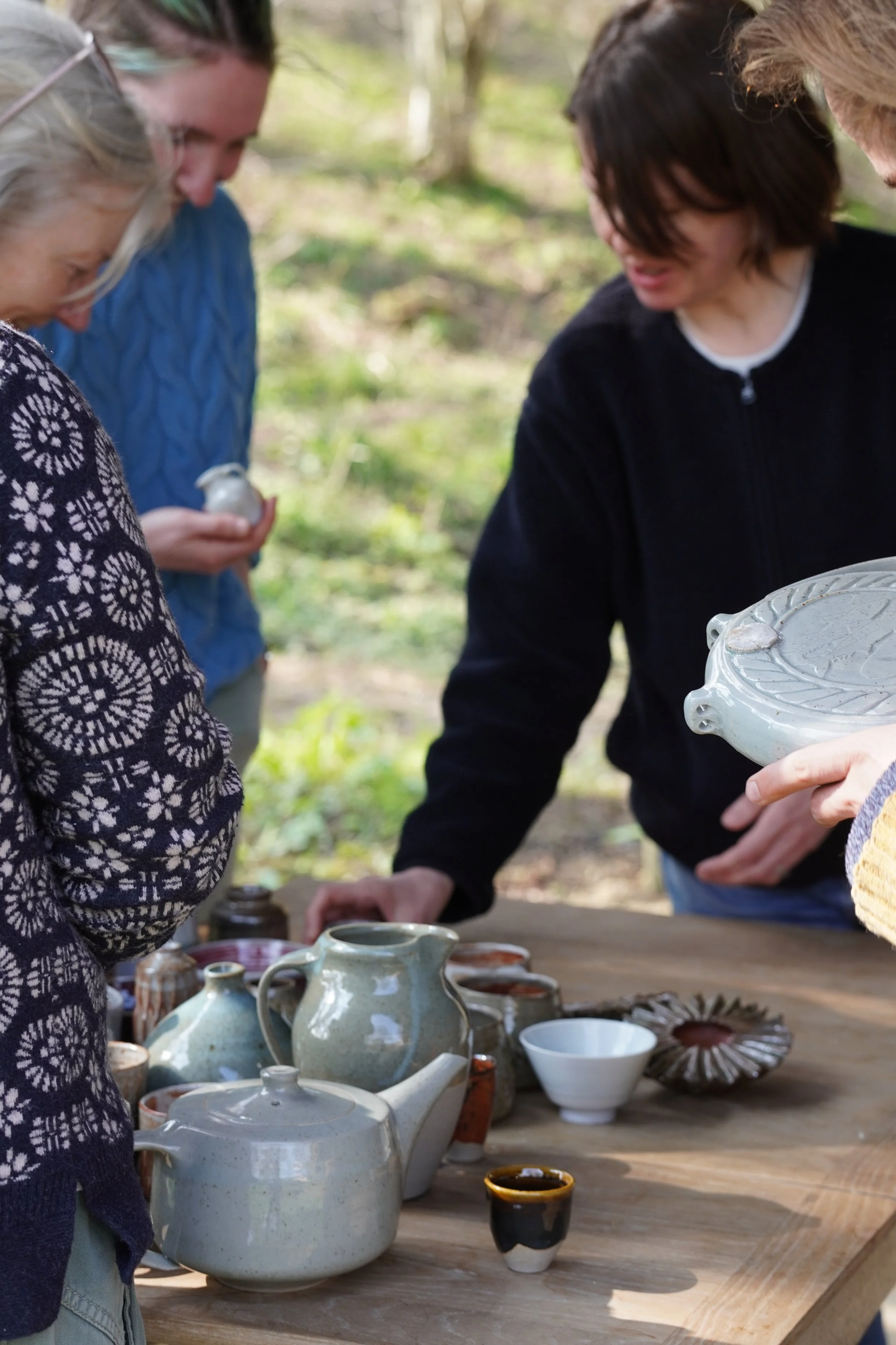 A group of people gathered around a wooden table outdoors, inspecting and possibly purchasing handmade pottery and ceramics including teapots, cups, and decorative bowls.