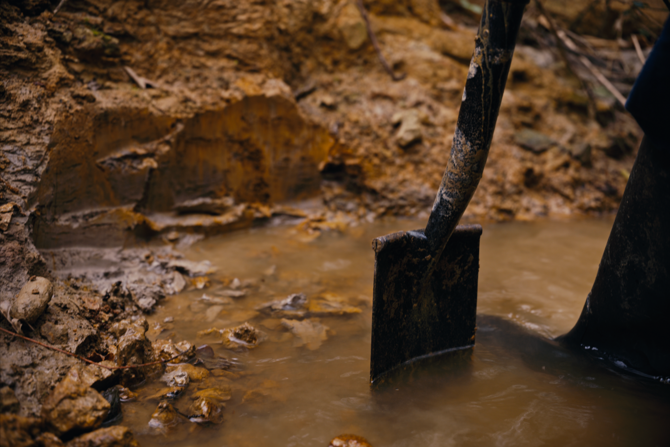 Close-up of muddy ground with a partial view of a shovel stuck in wet soil near a small muddy stream.