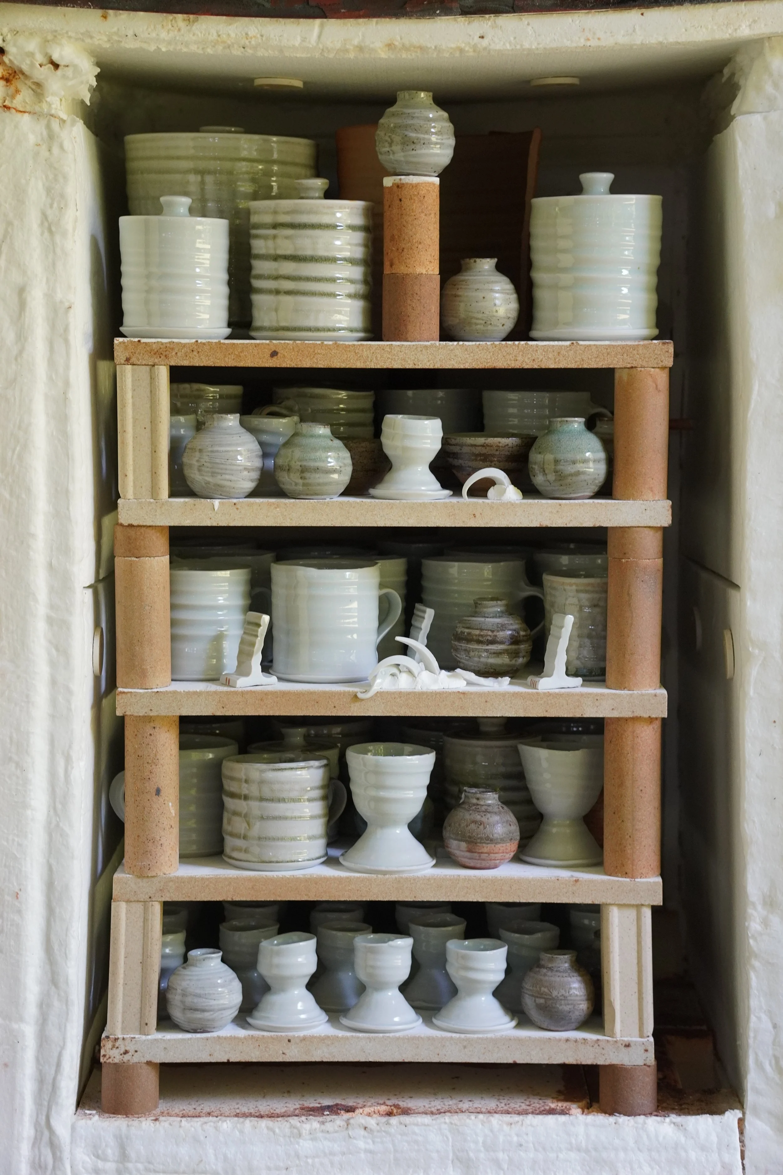 Shelf filled with various ceramic pottery pieces including vases, bowls, and cups, arranged on multiple tiers with some pieces having a striped or glazed pattern.