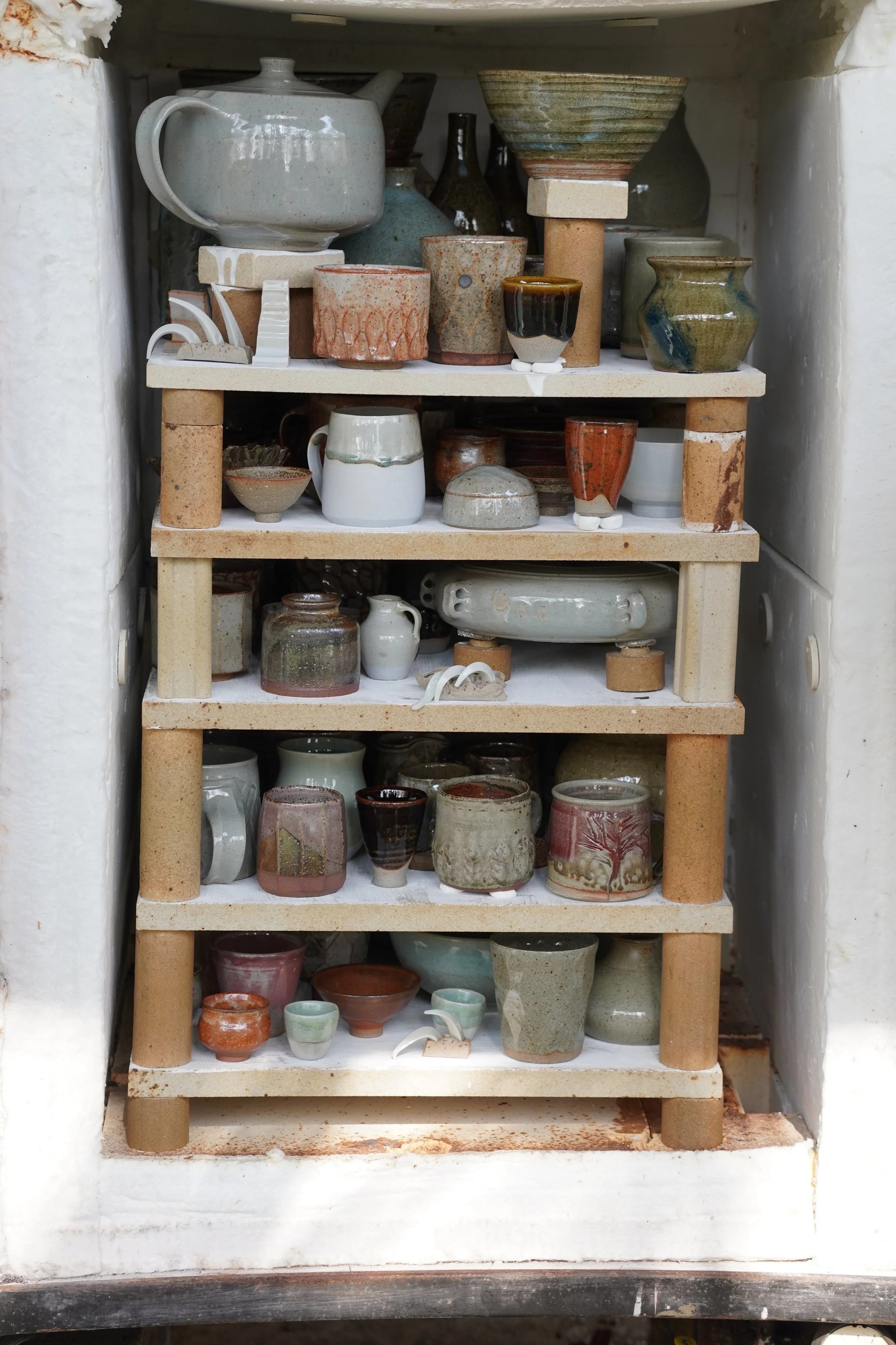 Shelf with various handmade ceramic cups, bowls, and vases in different shapes, sizes, and colors, placed in a recessed niche with white walls.