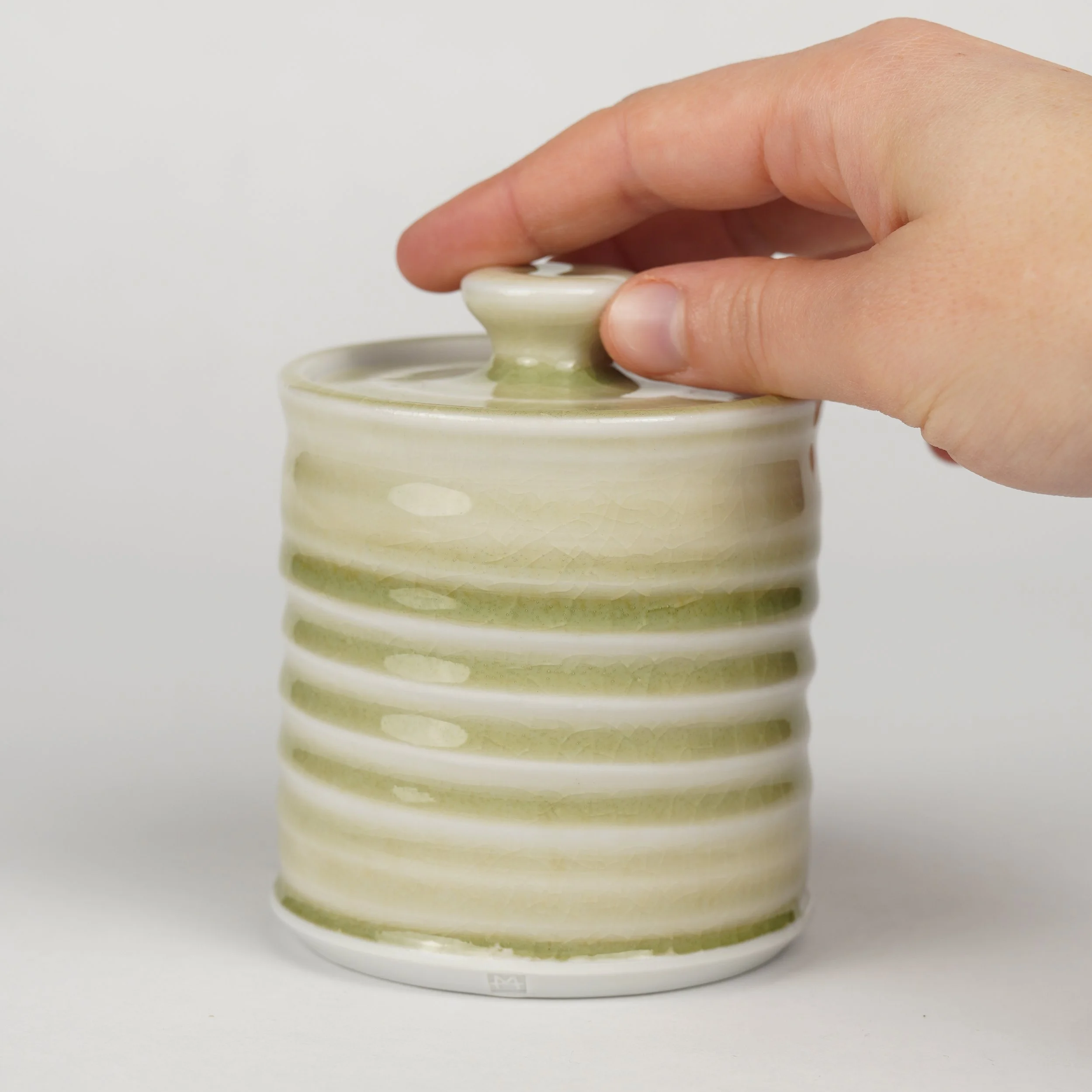 Close-up of hand opening a ceramic jar with a rounded lid, cream and green glaze, against a plain white background.