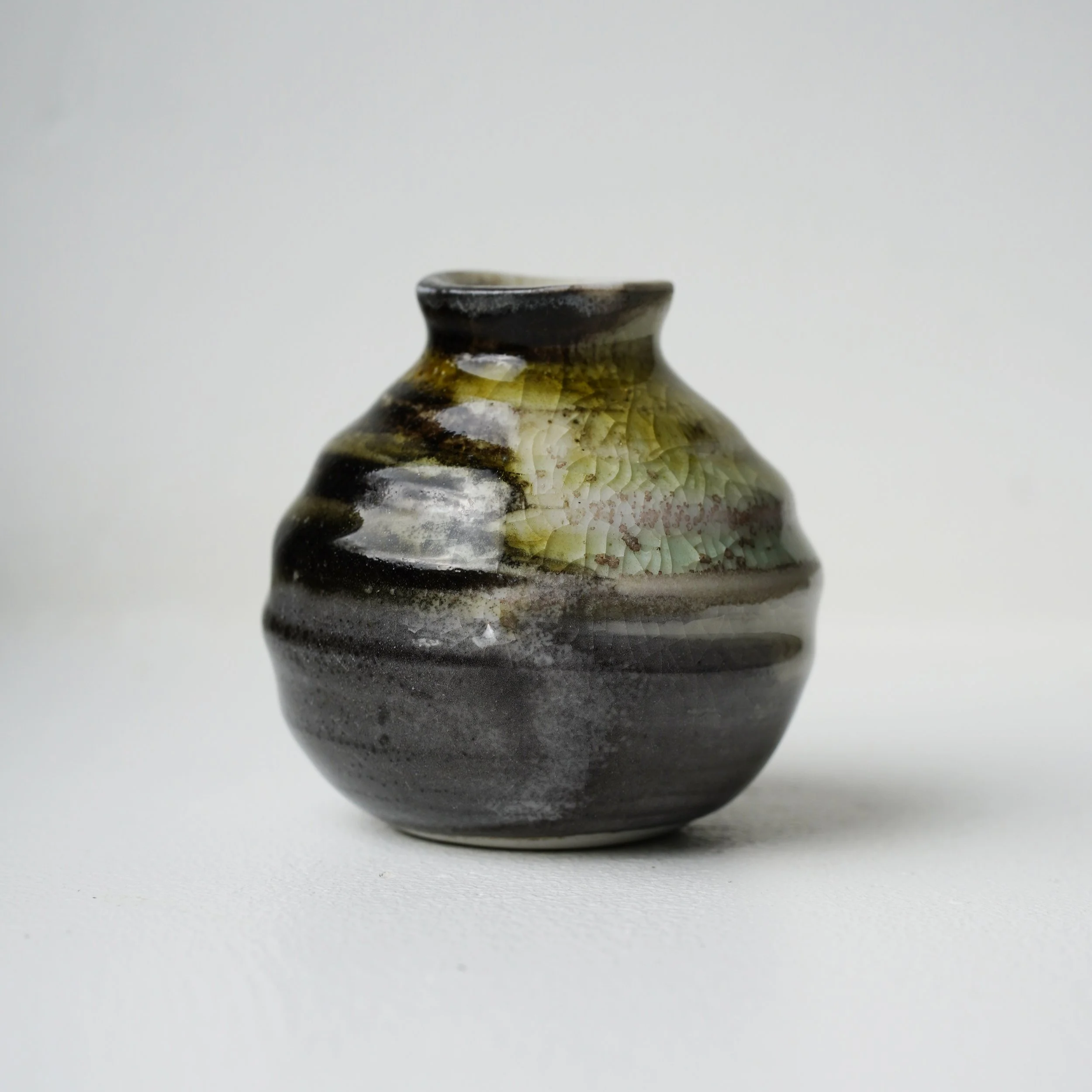 Close-up of a ceramic vase with a glossy, textured surface, displaying earthy tones of black, brown, green, and cream, set against a plain white background.
