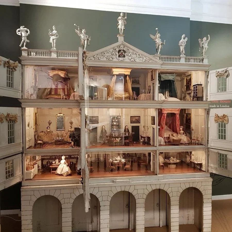 A detailed dollhouse with four floors, styled as a Victorian mansion. The interior is decorated with miniature furniture, including living rooms, bedrooms, a dining area, and hallway, with miniature dolls in period costumes. The top floor features a staircase and small statues on the roof. The dollhouse has windows and arches, and is set against a green wall.