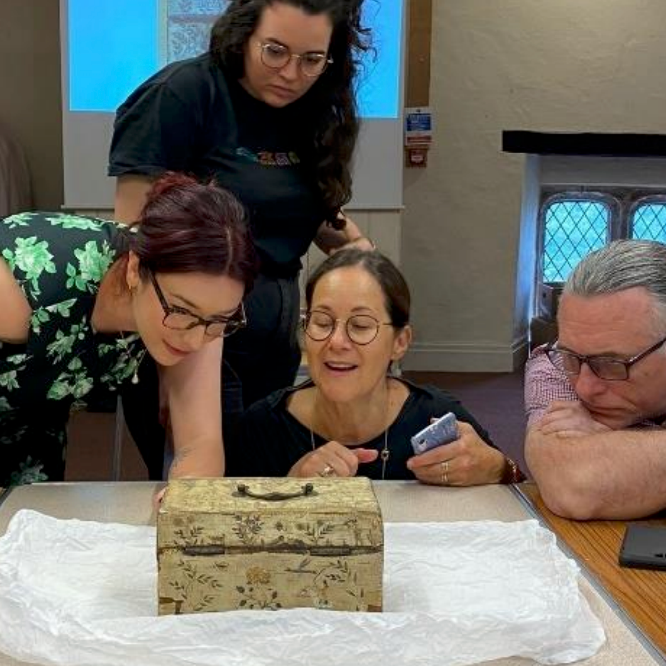 Group of four people gathered around a table, looking at a small decorative wooden box on white paper, with two women leaning over and two people sitting, one of whom is holding a smartphone.