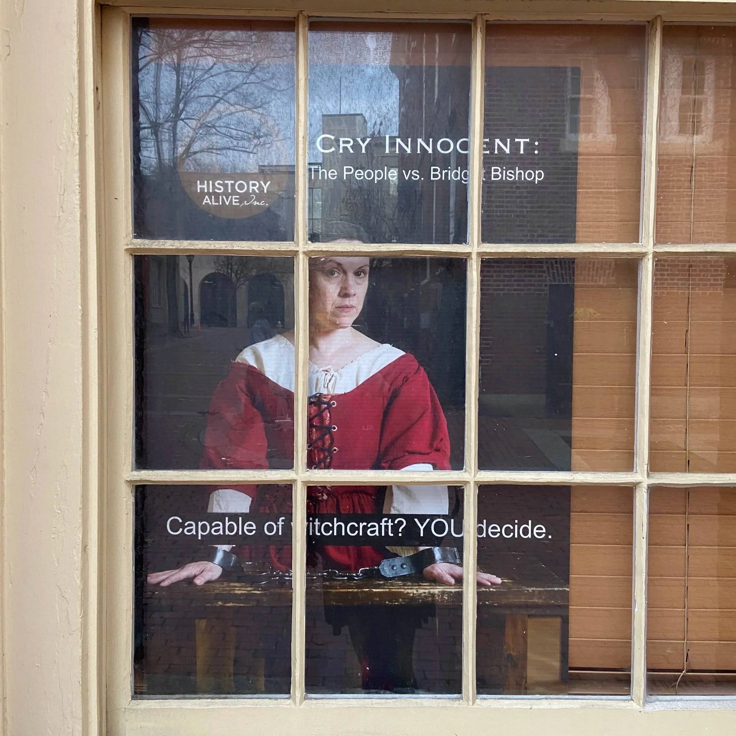 Window with a sign reading 'Cry Innocent: The People vs. Bridget Bishop' and 'History Alive, Inc.', reflecting trees and sky outside. A woman in a red dress is visible through the lower part of the window.