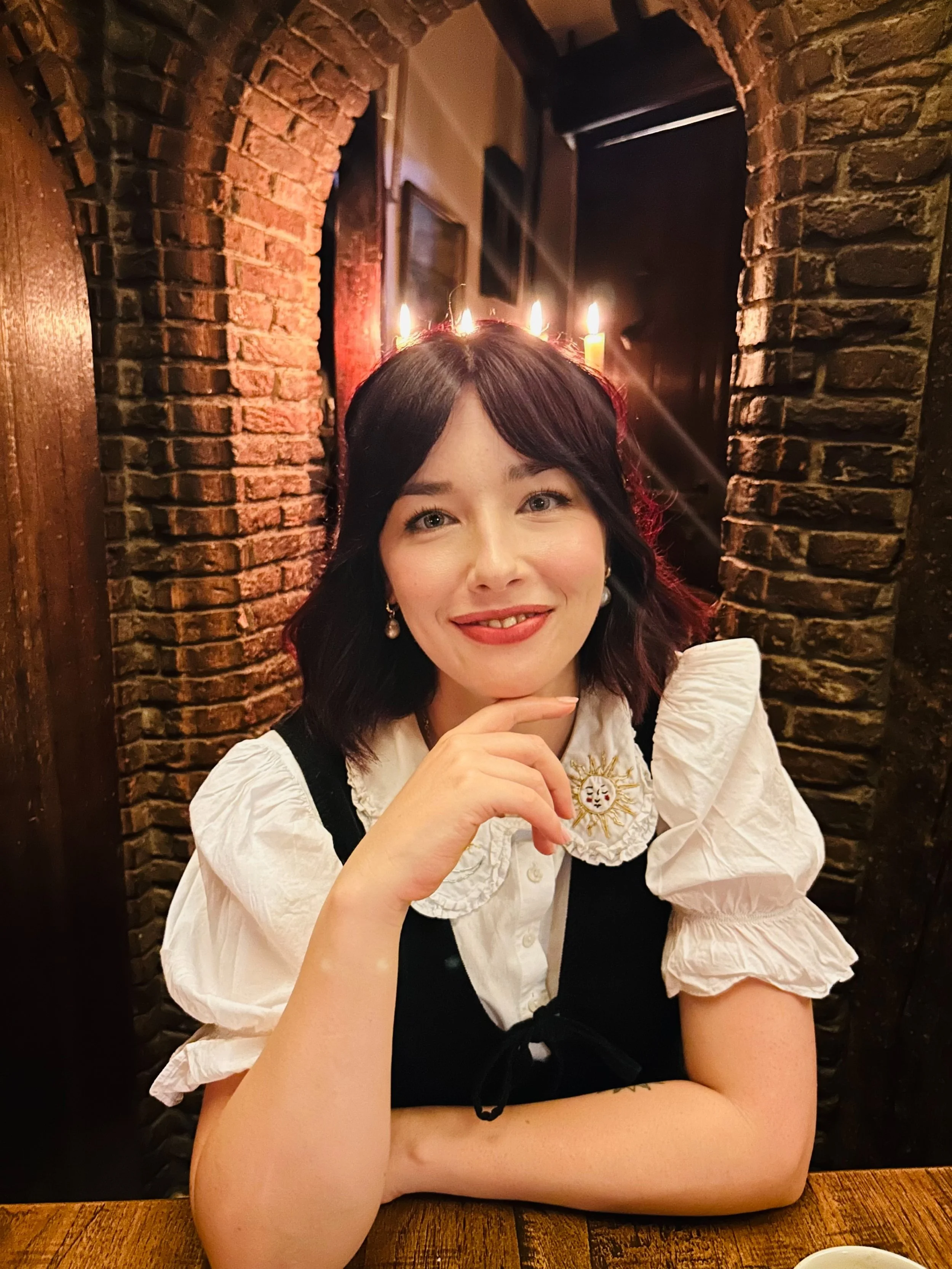 A woman sitting at a wooden table in a restaurant, smiling, wearing a white blouse with a decorative patch and pearl earrings, with a black bow on her sleeve. Behind her are brick walls and candles on the wall.