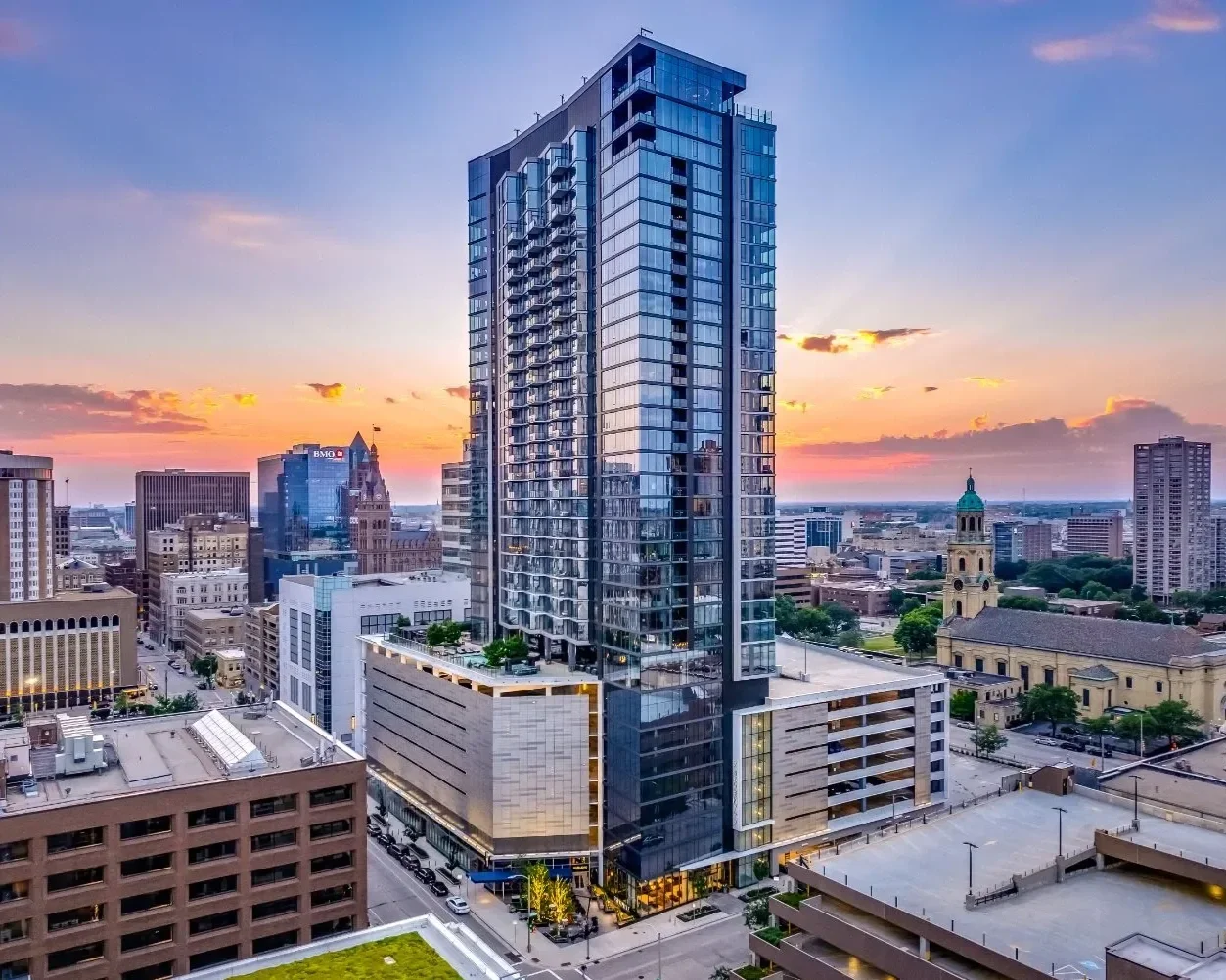 City skyline at sunset featuring a tall modern glass skyscraper surrounded by older buildings, with a church and sunset sky in the background.